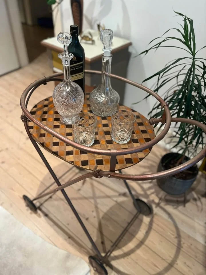 A vintage metal bar cart with a wooden top, holding two glass decanters and two glasses, situated in a room with hardwood floors, a potted plant, and a small table with various items in the background.
