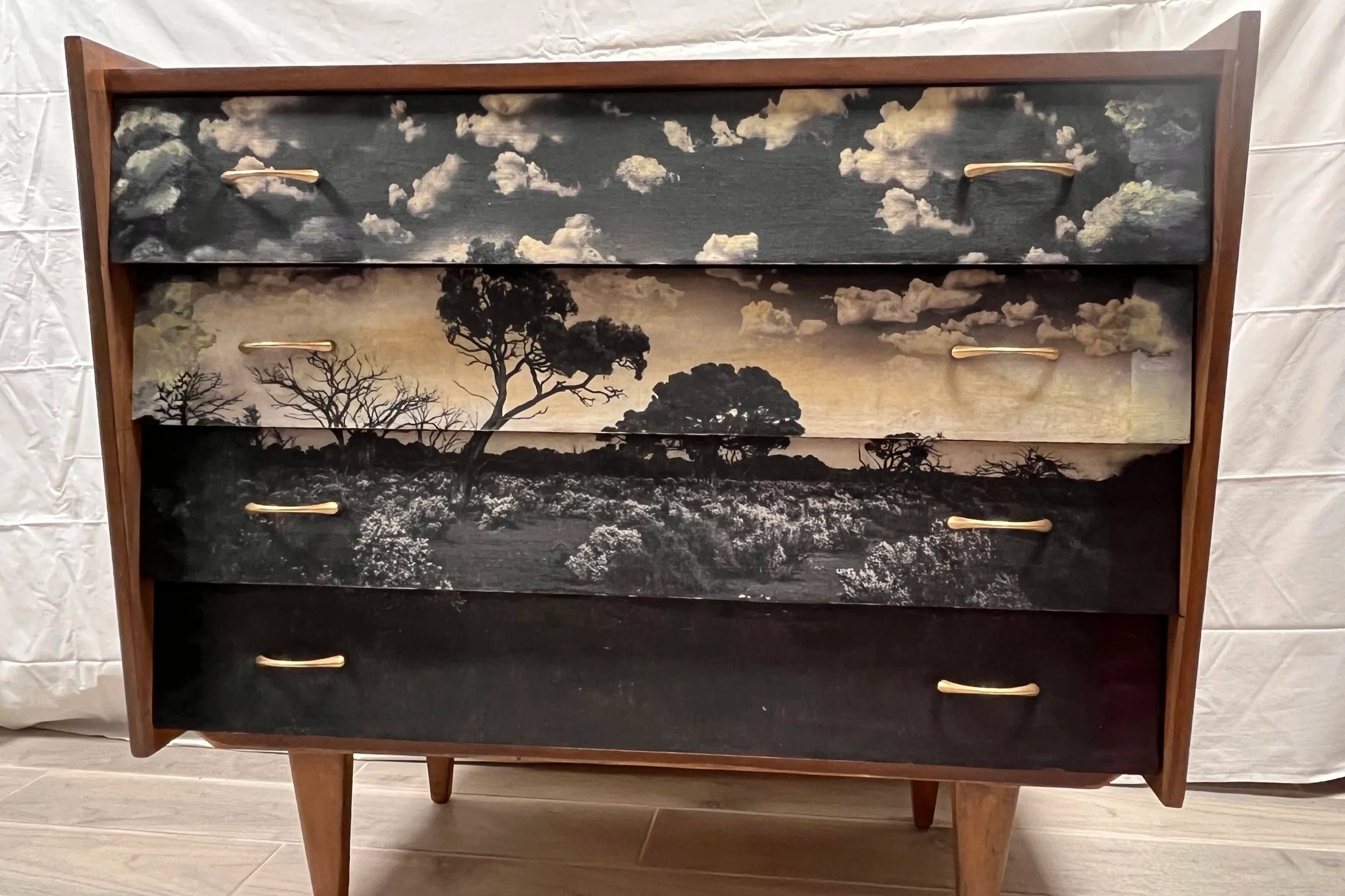 A wooden dresser with four drawers, each painted with a landscape scene of trees and clouds in shades of black, white, and sepia, with gold-colored handles.