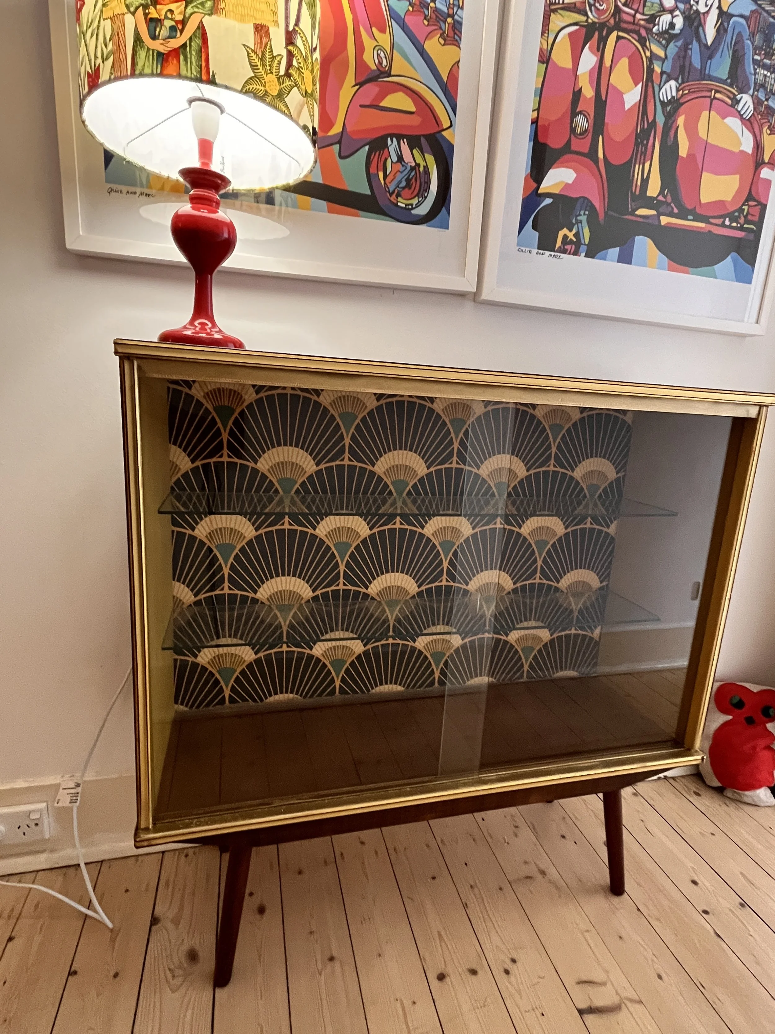 Mid-century modern display cabinet with glass doors, gold framing, and a patterned back panel. A red lamp with a colorful shade sits on top, and vibrant art prints are on the wall behind. Wooden floor visible.