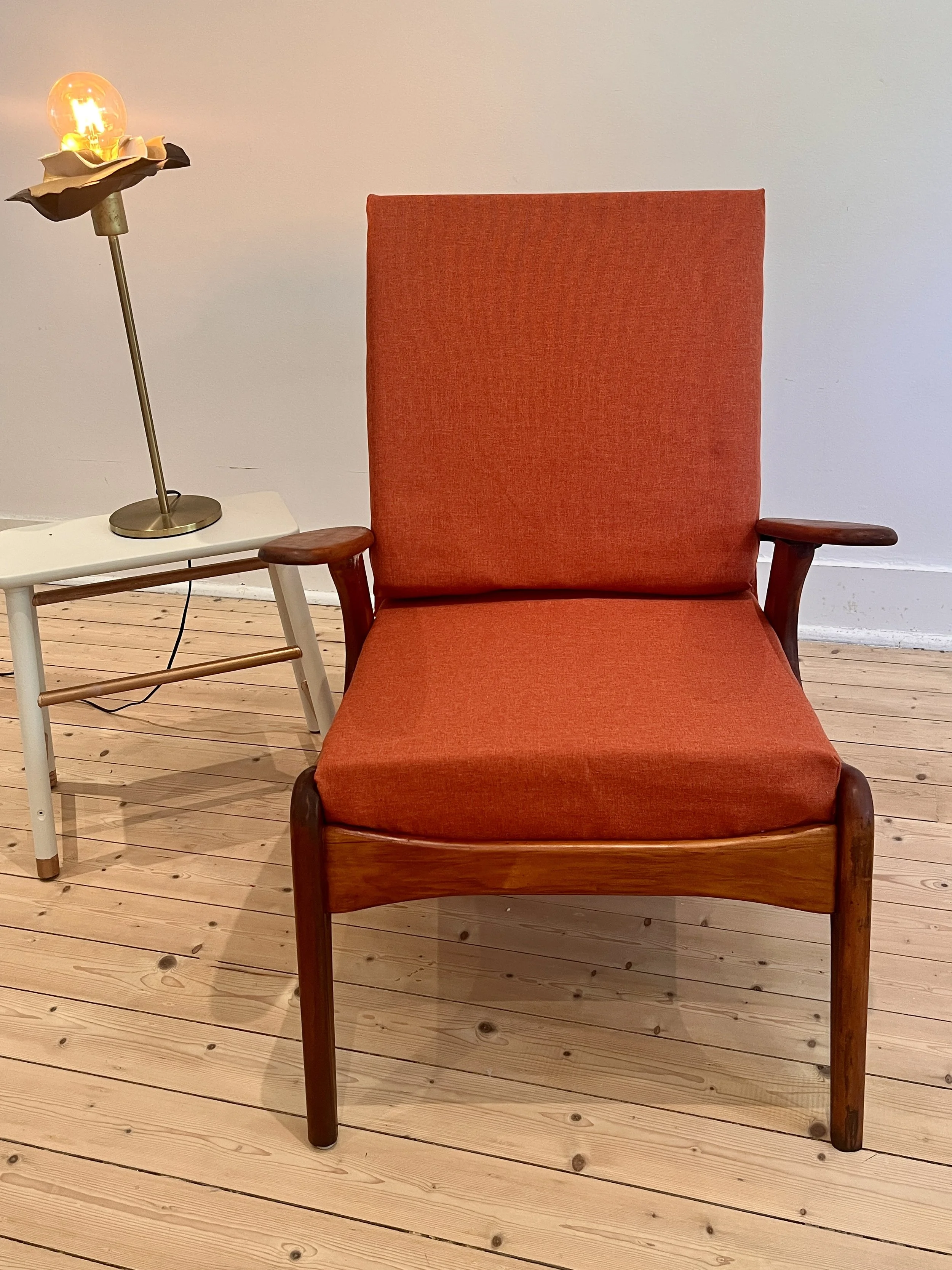 An orange upholstered armchair with wooden armrests and legs, positioned on a wooden floor against a plain white wall. To the left, there is a small white side table with a decorative lamp featuring a globe-style bulb and a flower-shaped shade.
