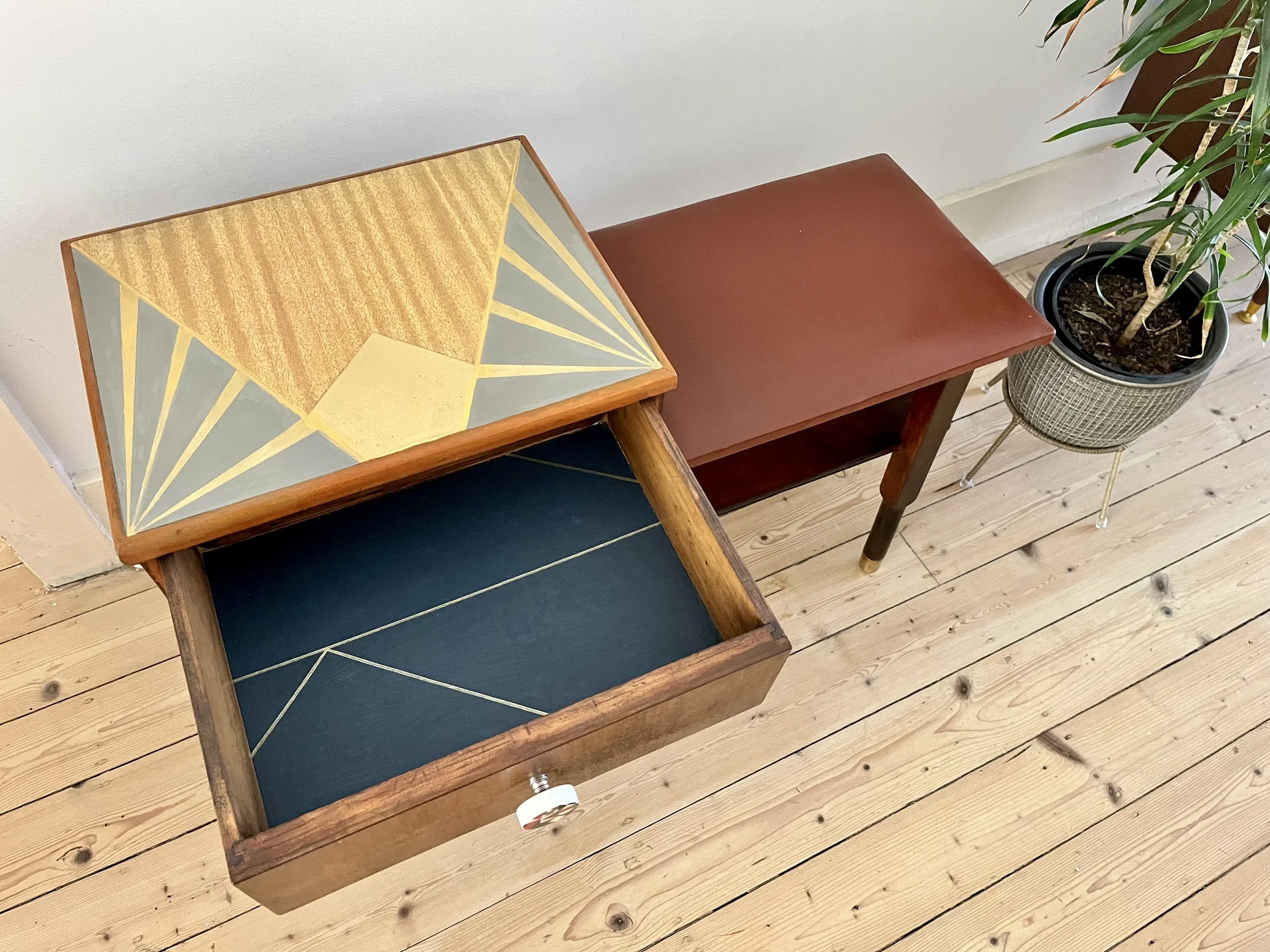 Open wooden drawer with geometric Art Deco design on top, black interior with gold lines, next to a brown table and a potted plant on a light wooden floor.
