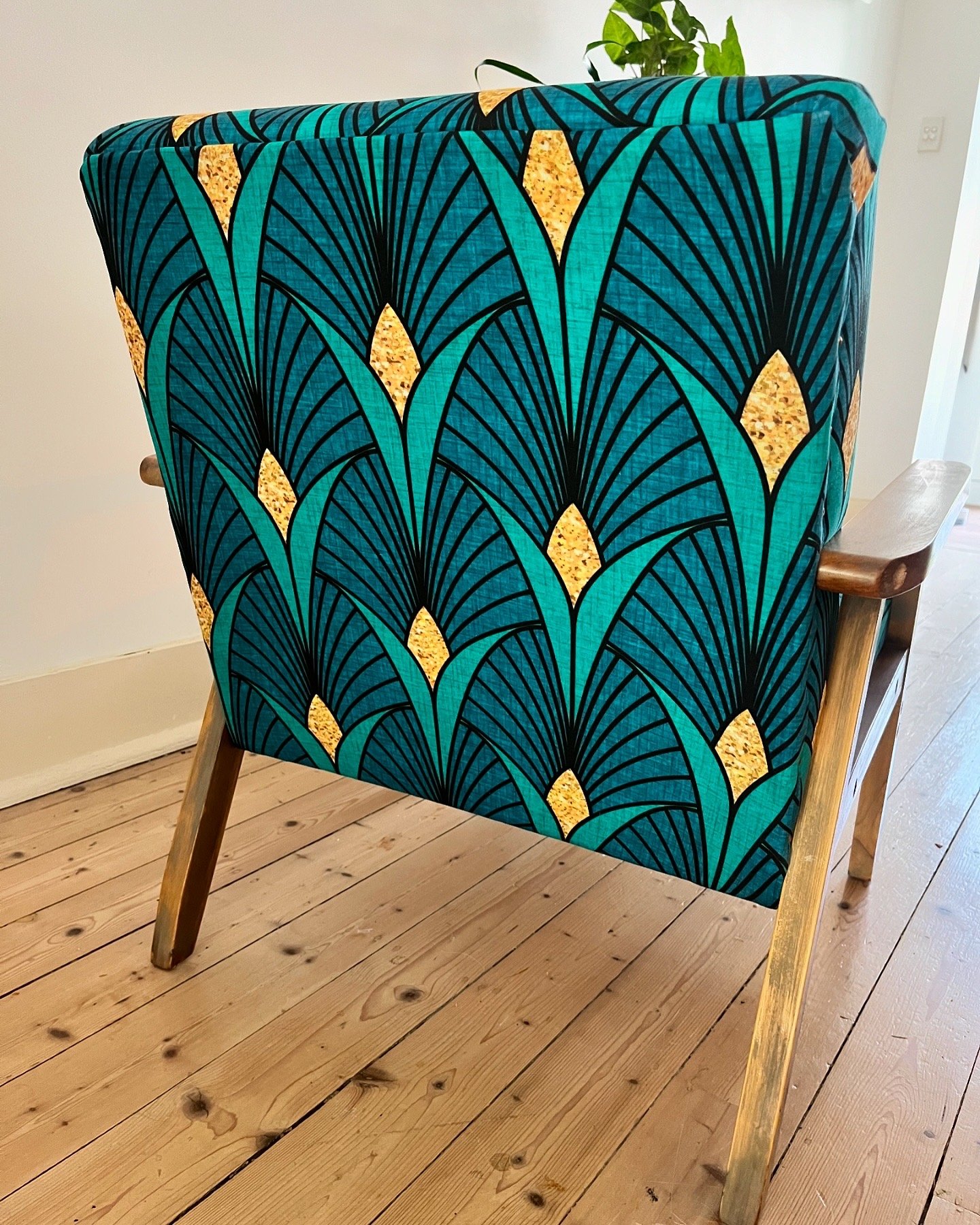 Wooden chair with a vibrant teal, black, and gold geometric patterned fabric upholstery, set on a light wooden floor with a small potted plant partially visible.