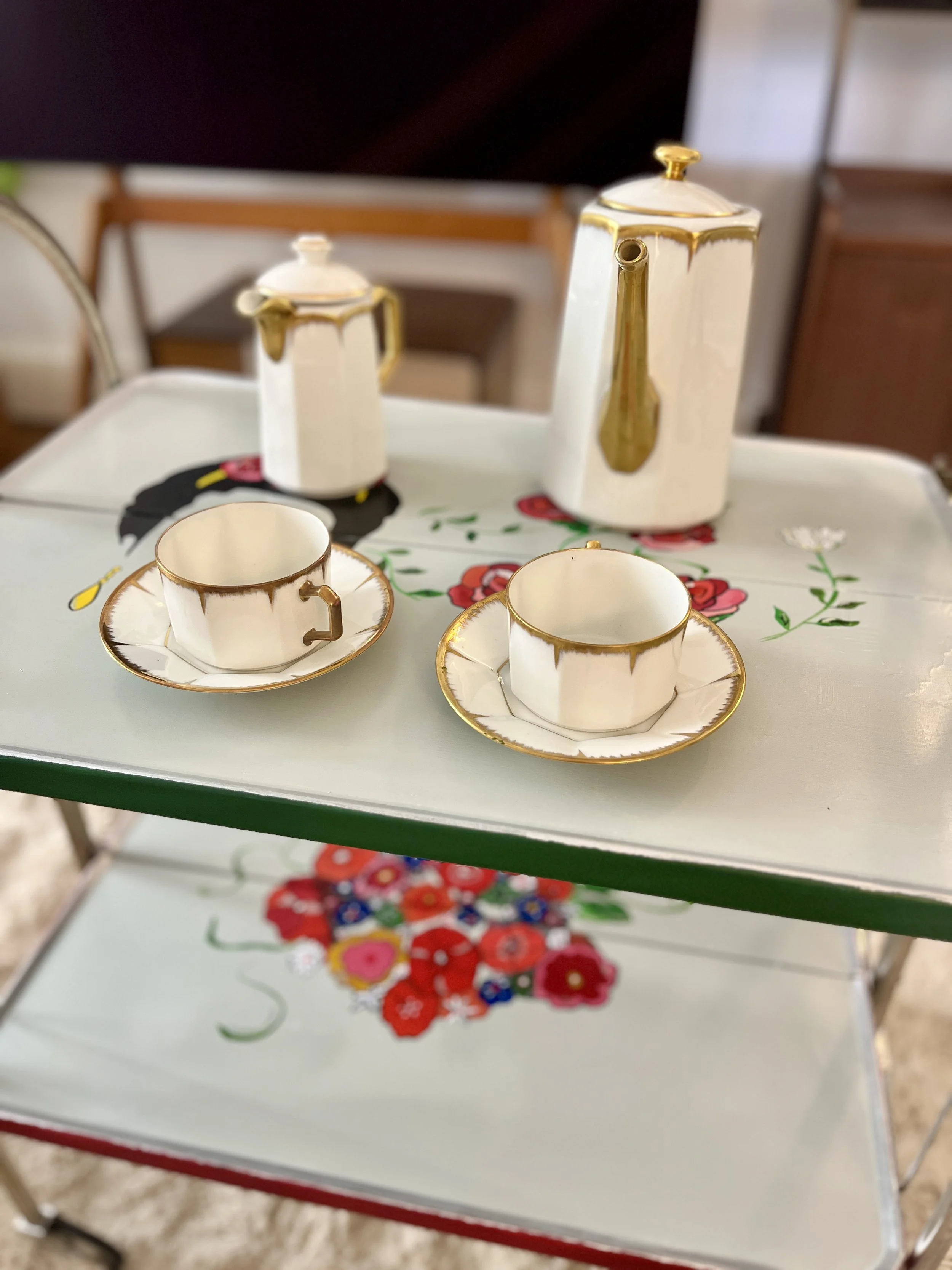 A vintage tea set with white cups and saucers decorated with gold trim, placed on a decorated table with floral designs. There are two teapots and a small milk jug in the background.
