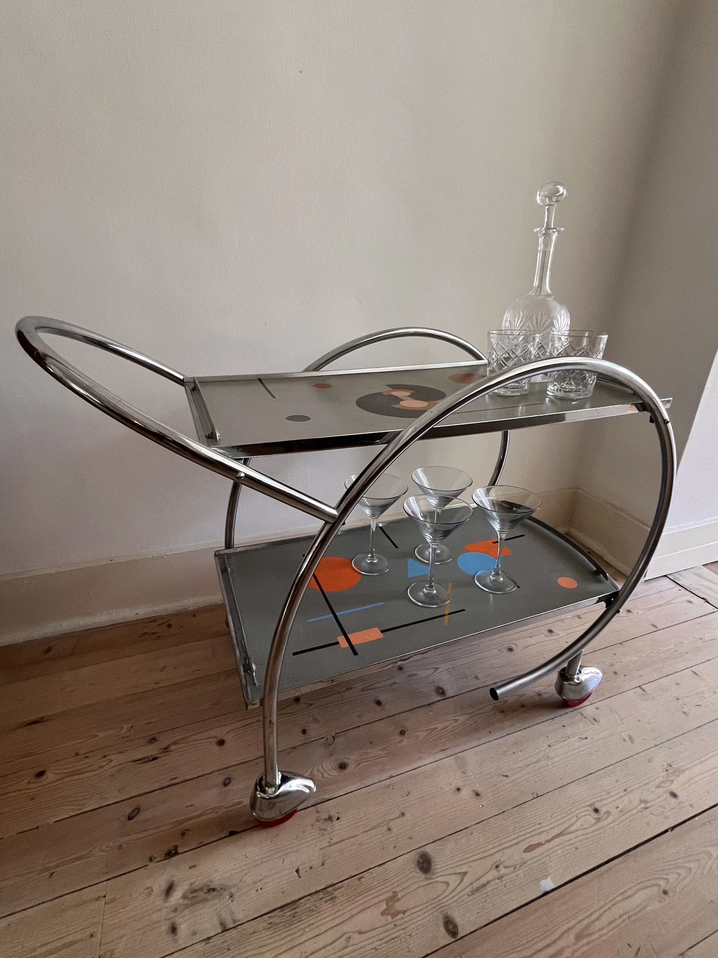 A modern, chrome bar cart with two glass shelves on wheels. The top shelf holds a clear glass decanter and two glasses. The bottom shelf features three martini glasses and colorful abstract geometric designs. The cart is placed on a wooden floor agai