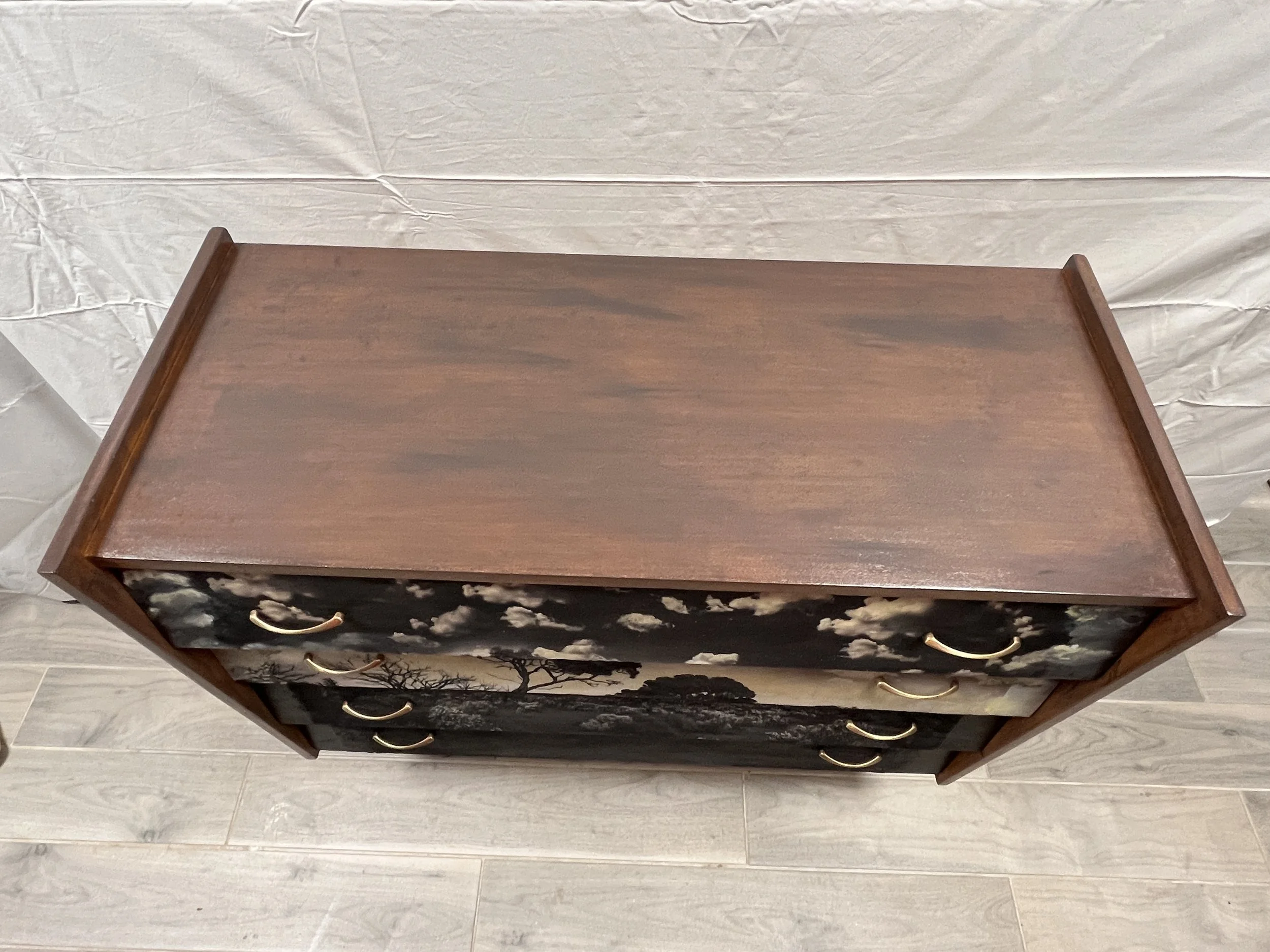 A wooden dresser with three drawers decorated with landscape painting and brass handles, placed on a light-colored wood floor against a neutral backdrop.