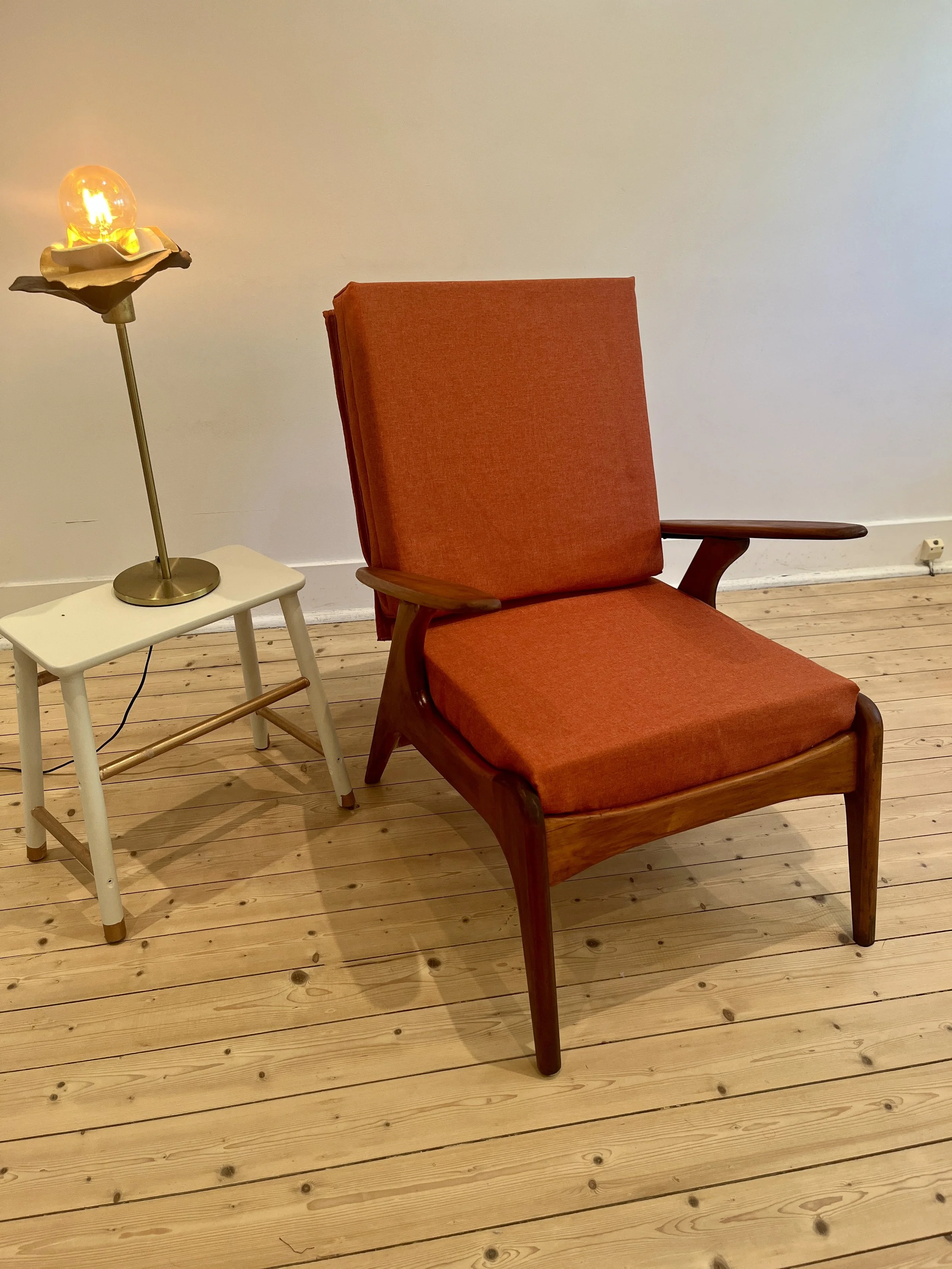 An orange upholstered armchair with wooden arms and legs, placed on a wooden floor. To its left is a white side table with a modern lamp featuring an Edison bulb shaped light bulb and a flower-shaped lampshade.