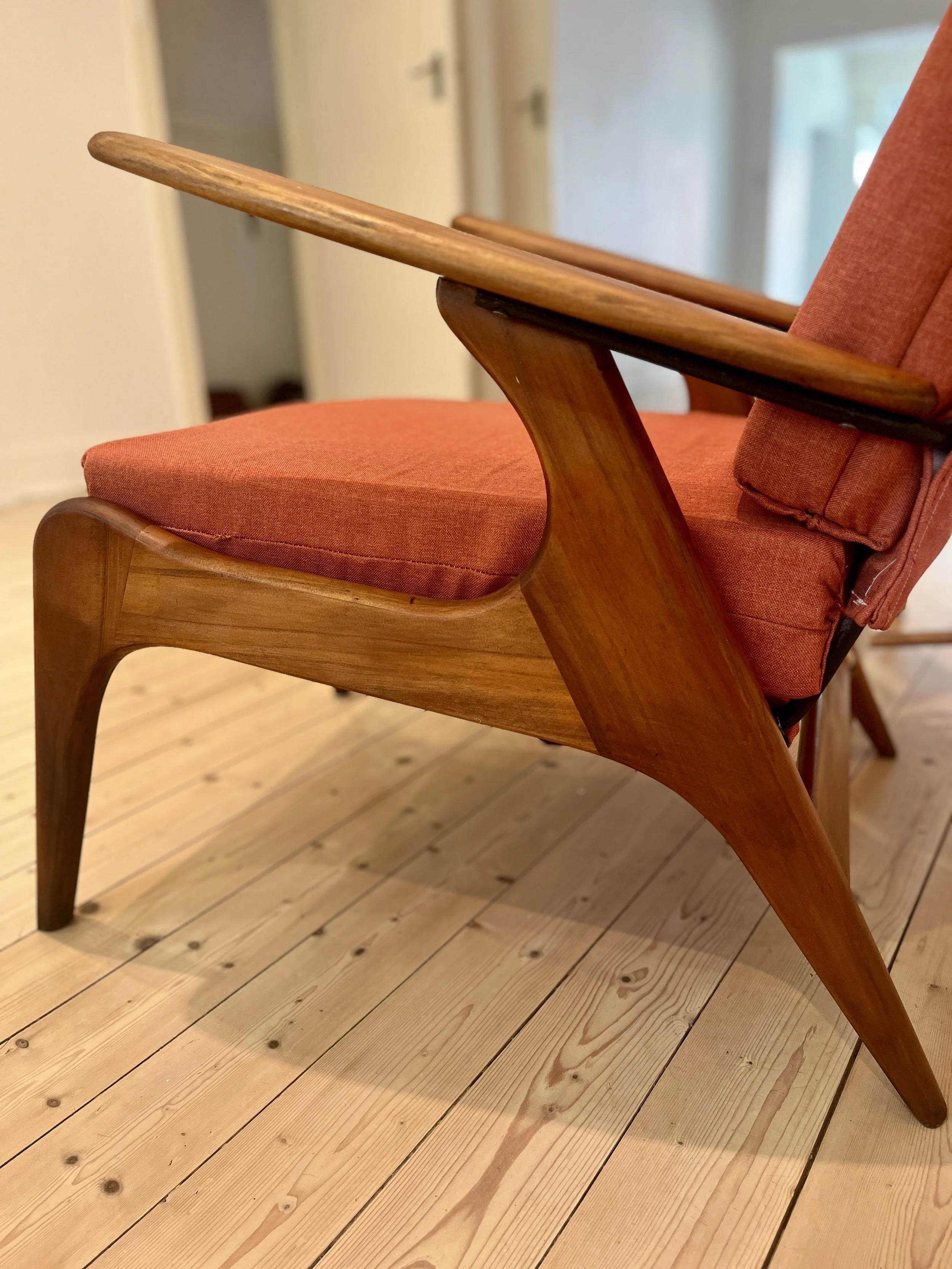 A mid-century modern wooden armchair with a reddish upholstered seat and backrest, placed on a light wood floor in a room with white walls and doors.