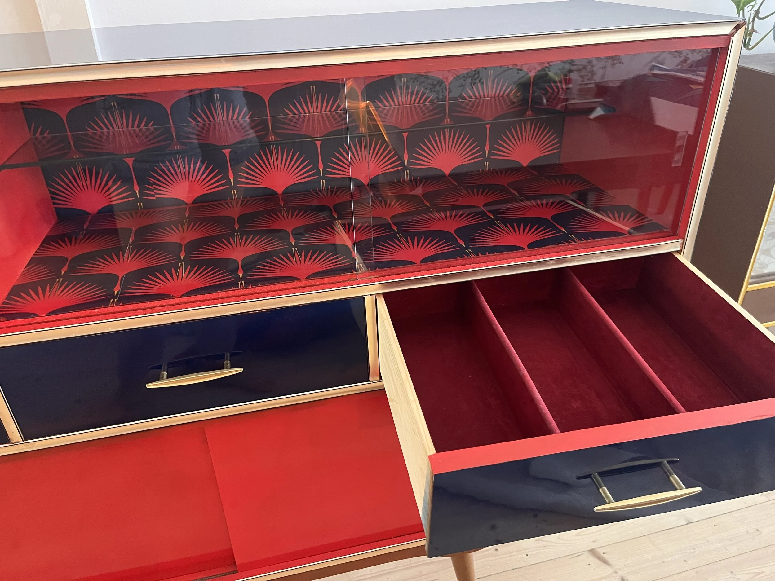A mid-century modern cabinet with glass doors displaying red and black patterned interiors, open drawers, and a gold handle.