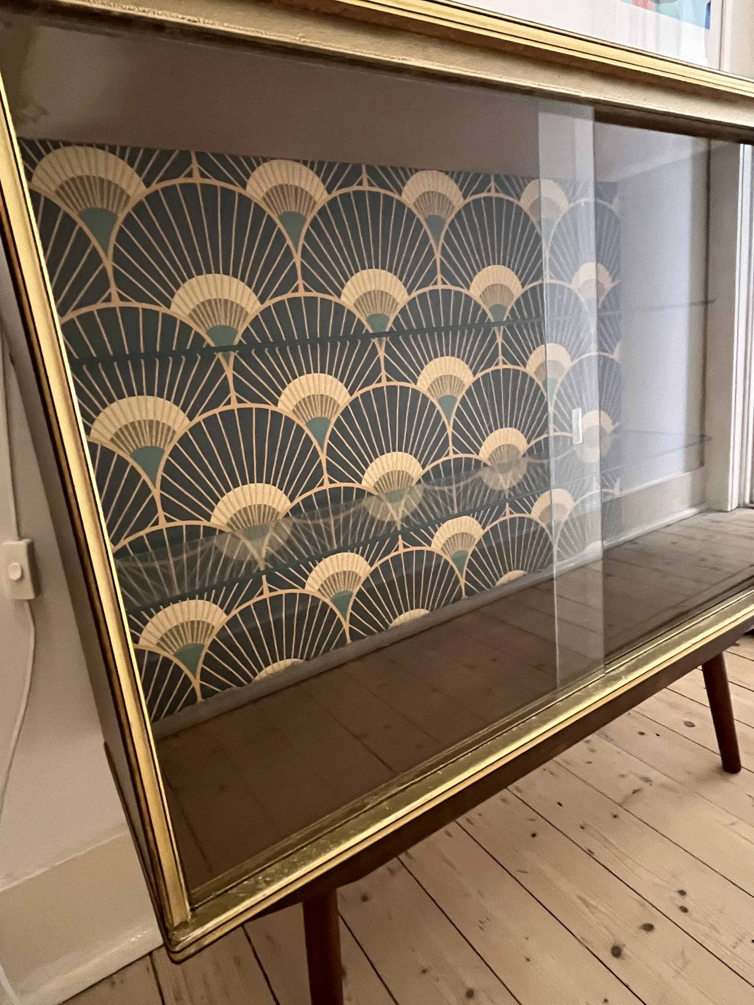 Glass display case with Art Deco wallpaper background, featuring geometric fan patterns in gold and teal.