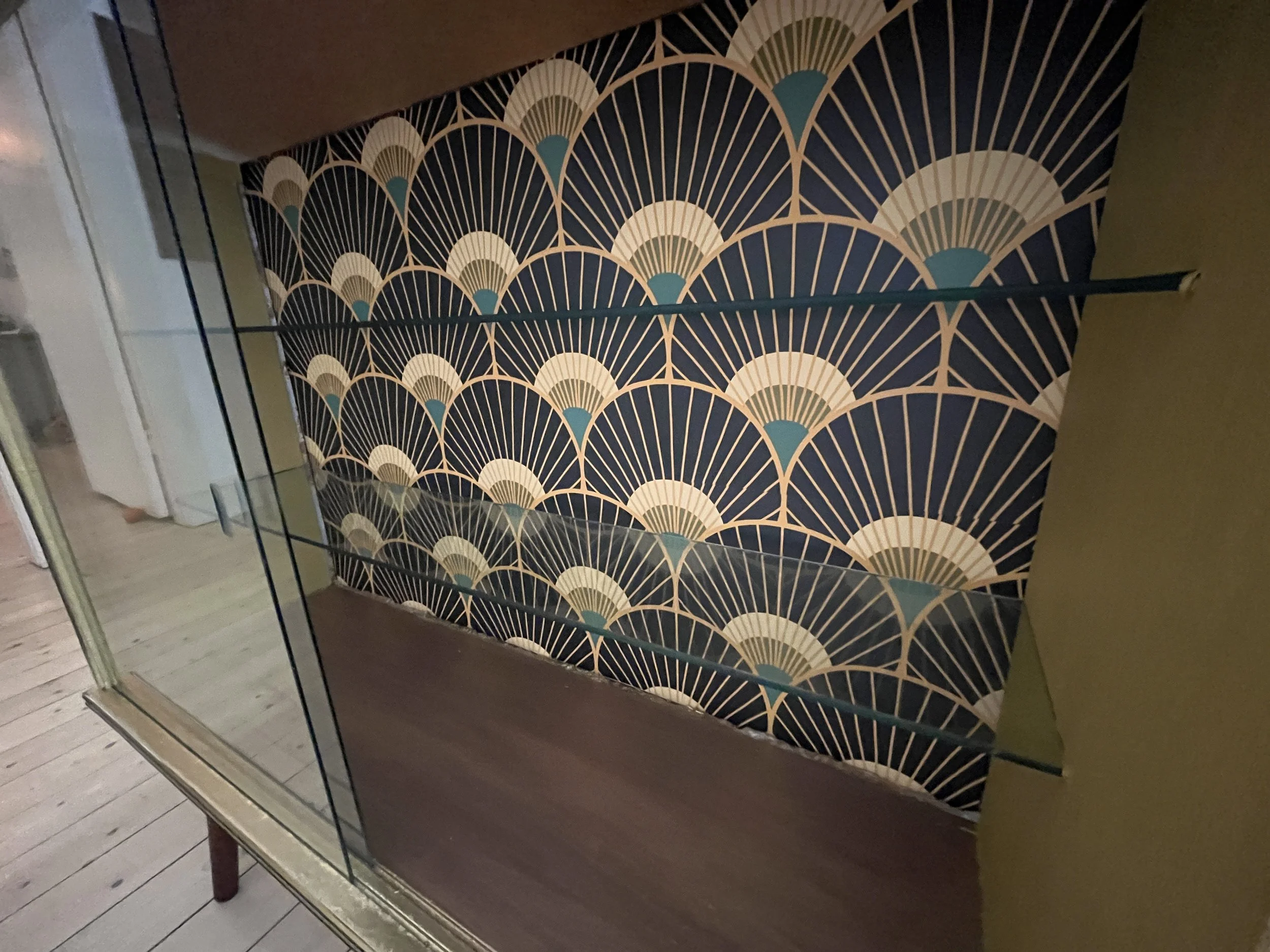Glass display cabinet with wooden base and art deco patterned wallpaper background featuring fan shapes in gold, teal, and white.