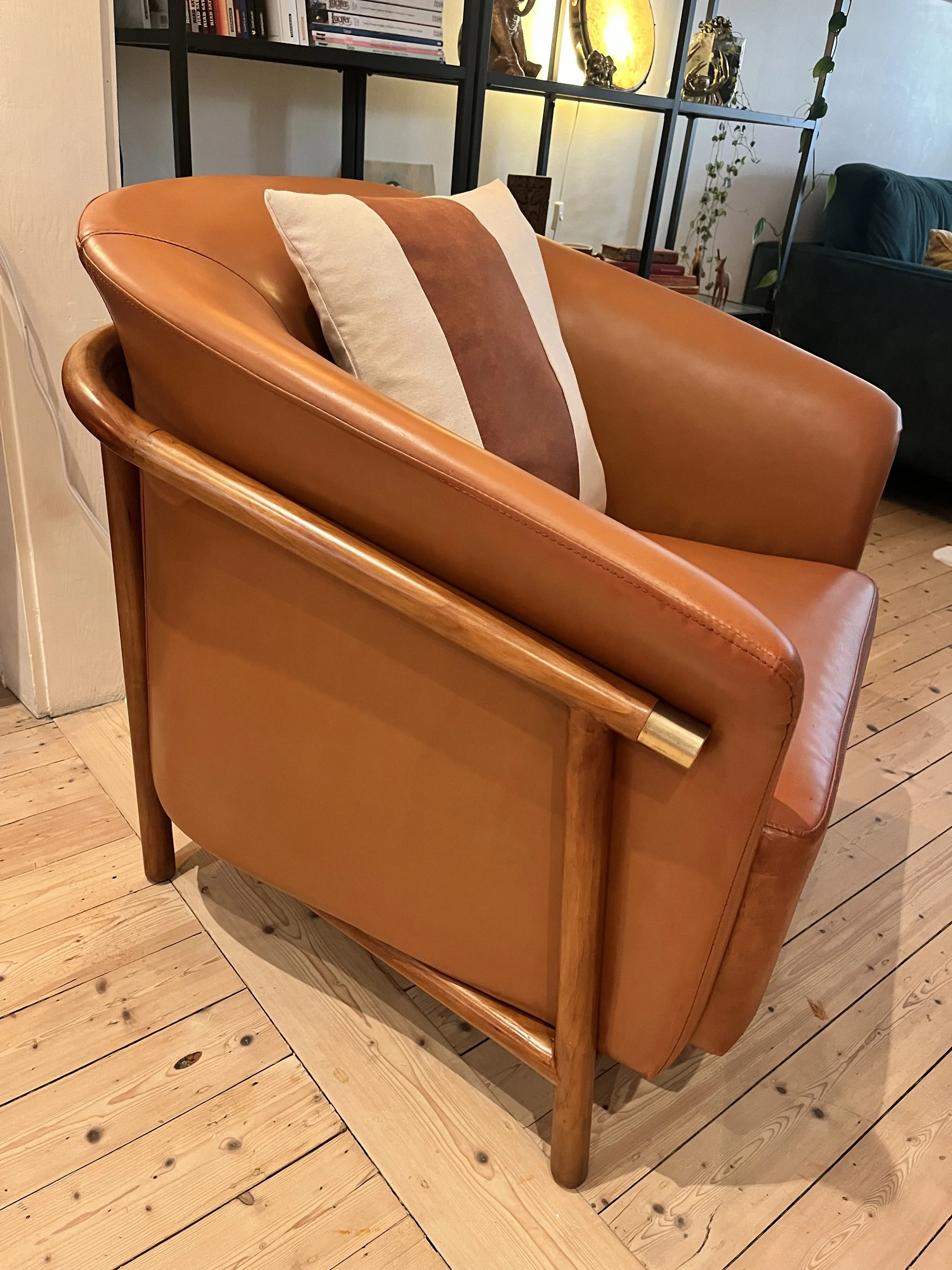 Modern leather armchair with wooden frame and a striped cushion on a wooden floor, in a cozy room setting with shelves and decorations in the background.
