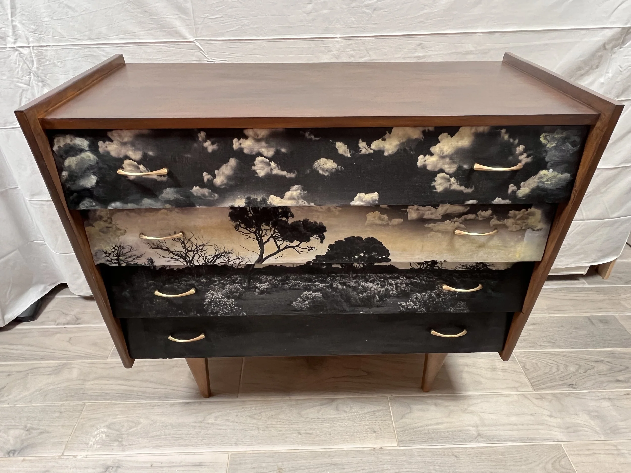 Four-drawer wooden dresser with sunset landscape painted on the front, featuring trees and clouds, against a light-colored floor and background.