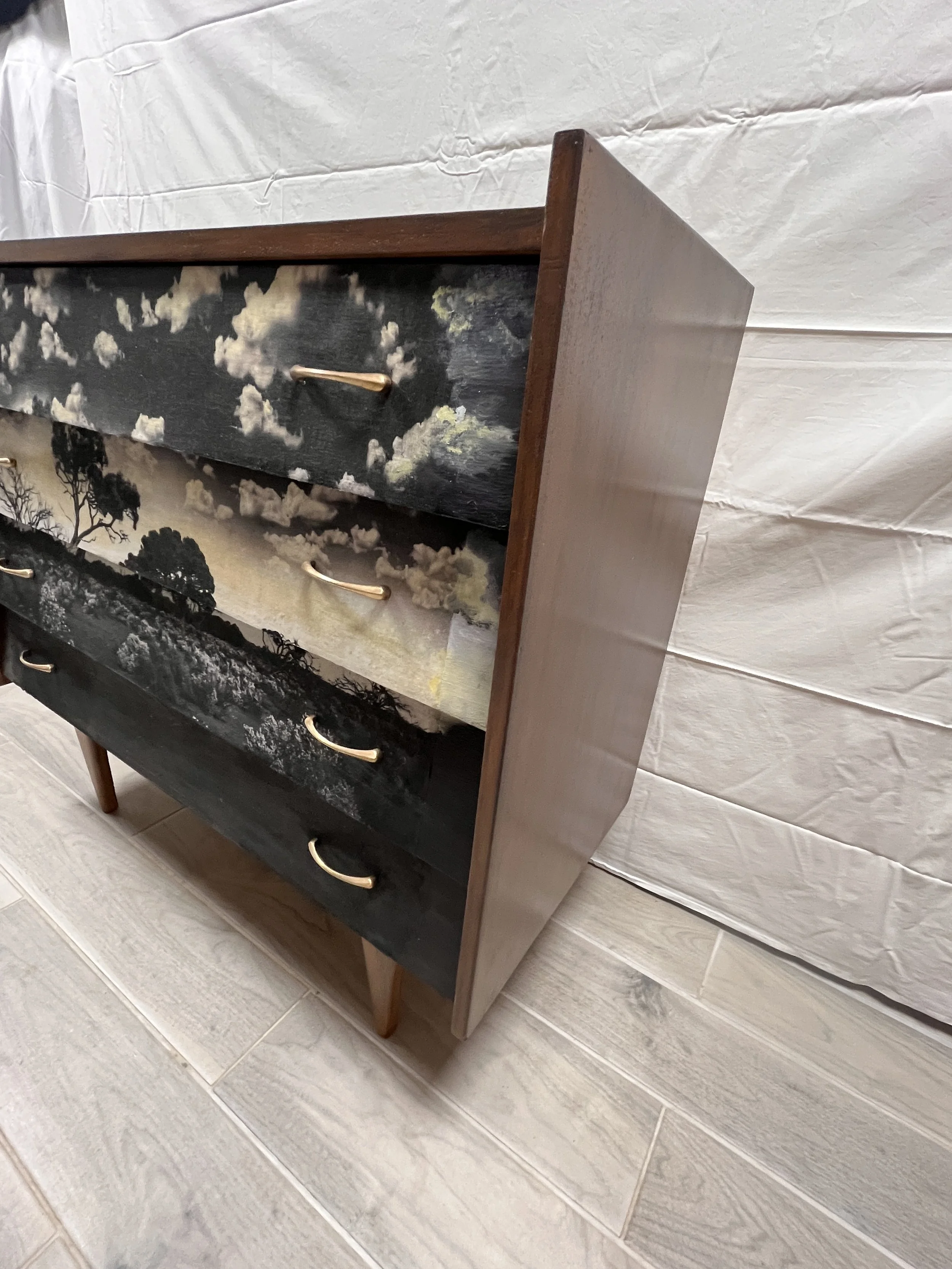 A wooden dresser with five drawers, each featuring a different landscape painting, including clouds, trees, and water, with curved brass handles.