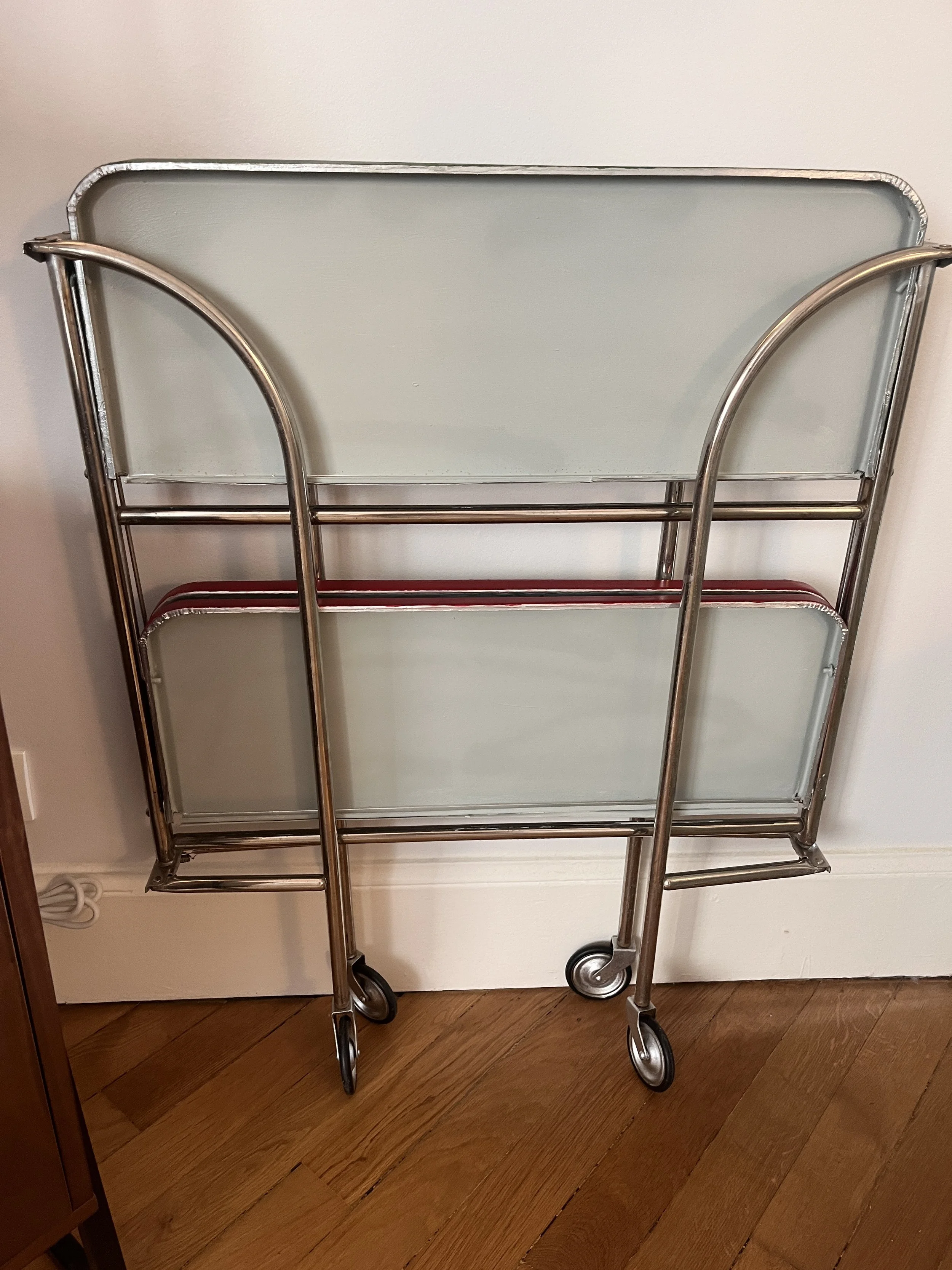 A vintage metal and glass rolling cart with two shelves, placed against a wall on a wooden floor.