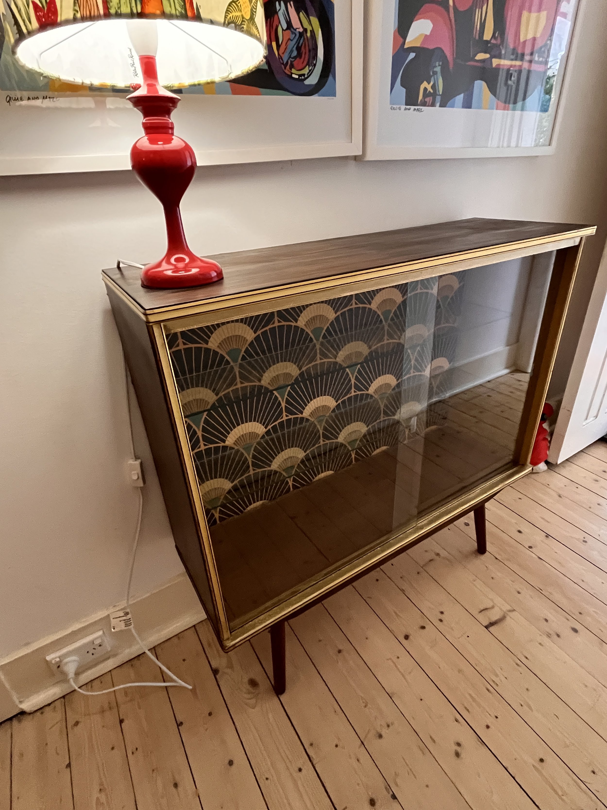 Vintage wooden cabinet with glass door, retro wallpaper inside, red lamp on top, hanging artwork in background.