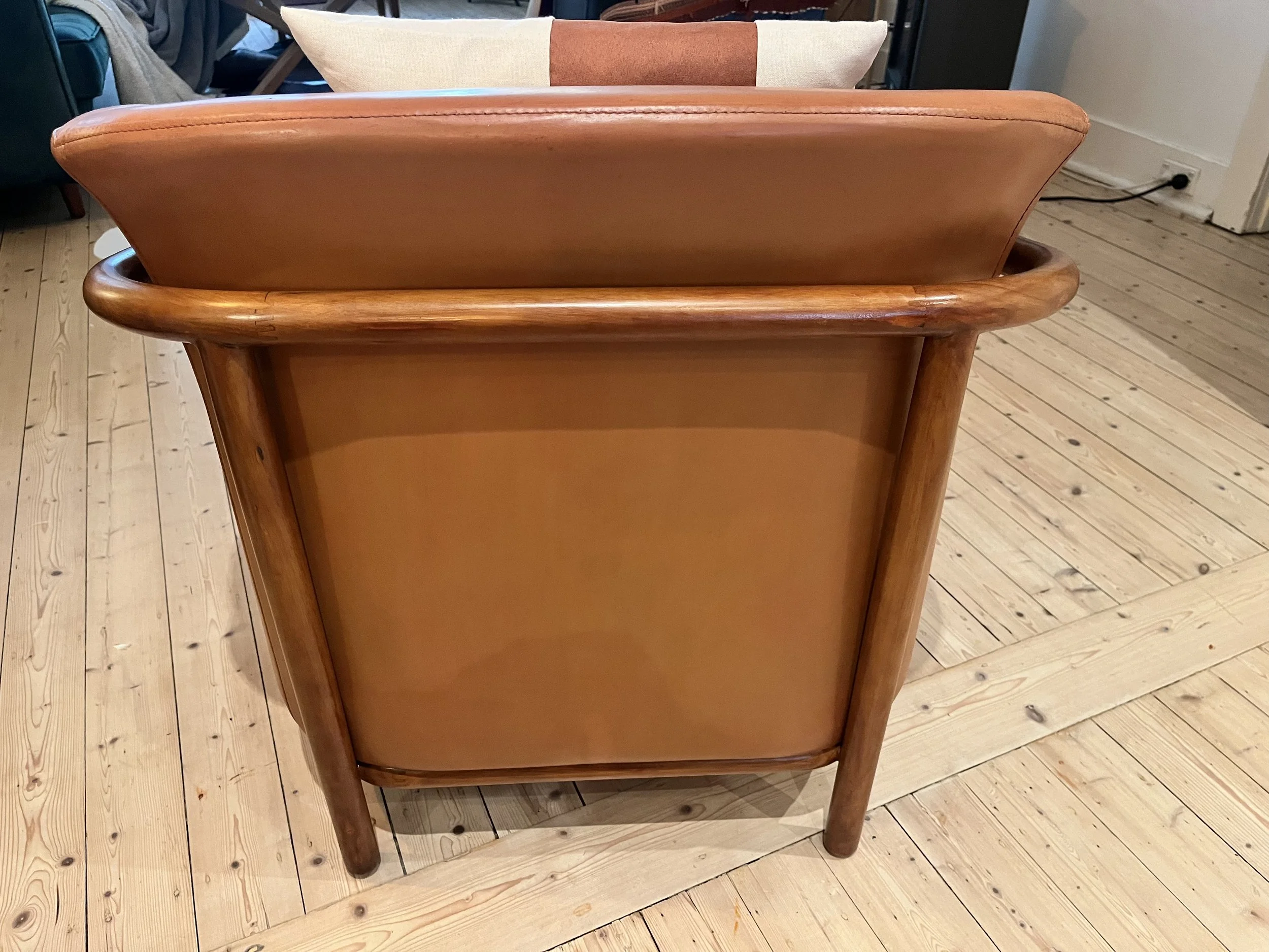 Back view of a brown leather armchair with wooden legs and frame on a wooden floor.