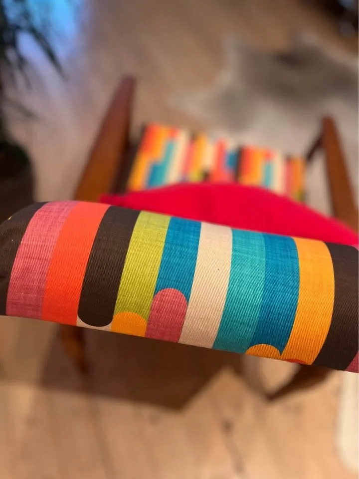 Close-up of a colorful striped fabric with a black background, draped over a surface, with a blurred wooden chair and a colorful striped object in the background.