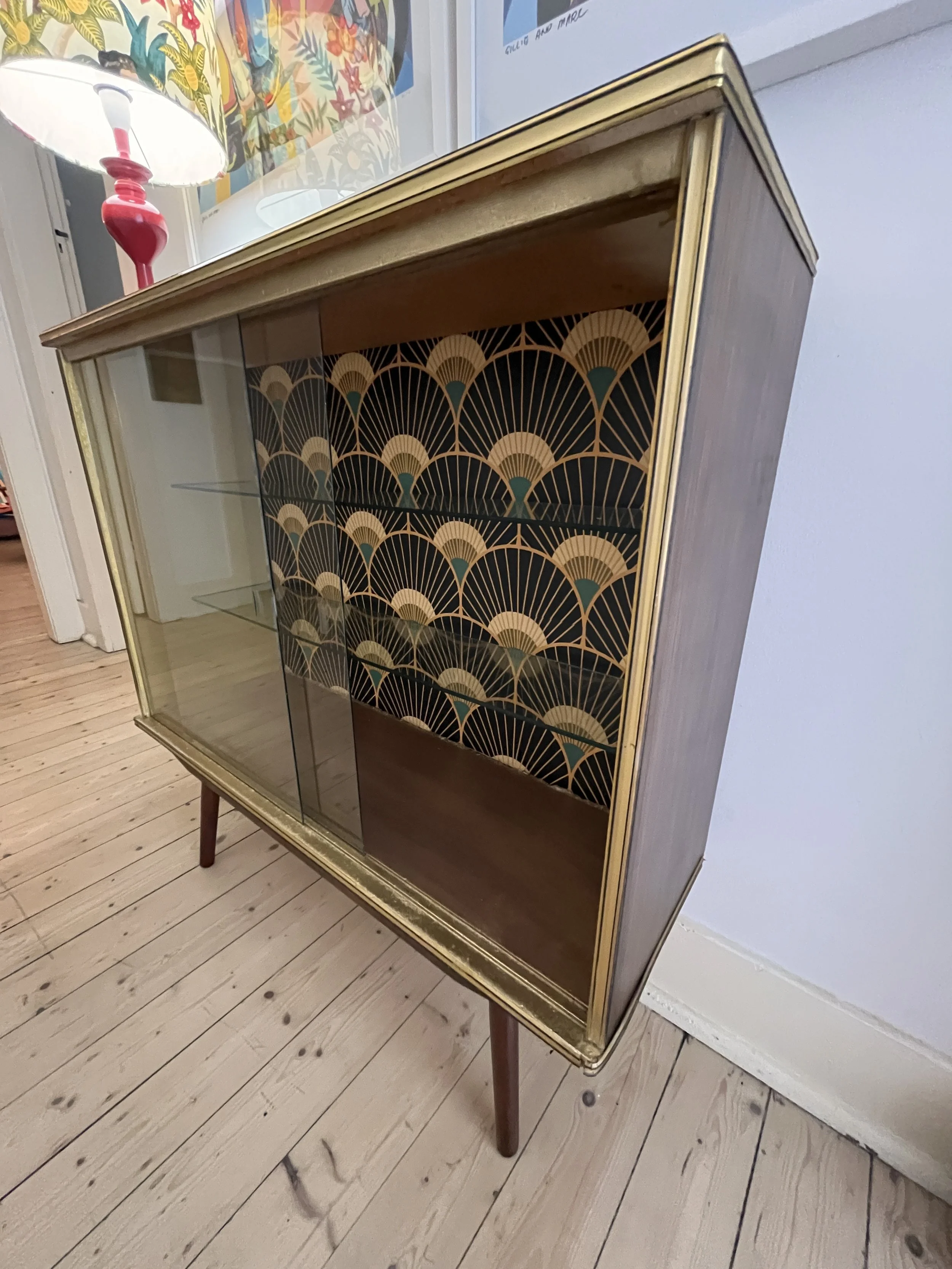 Vintage wooden cabinet with glass doors and patterned interior design, standing on a wooden floor next to a colorful lamp.