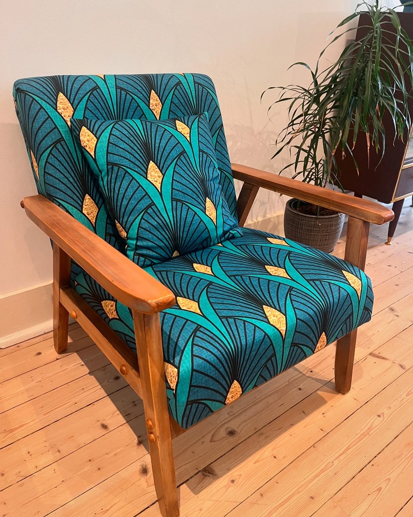 Armchair with colorful patterned upholstery and wooden frame, placed on a wooden floor, next to a potted plant.