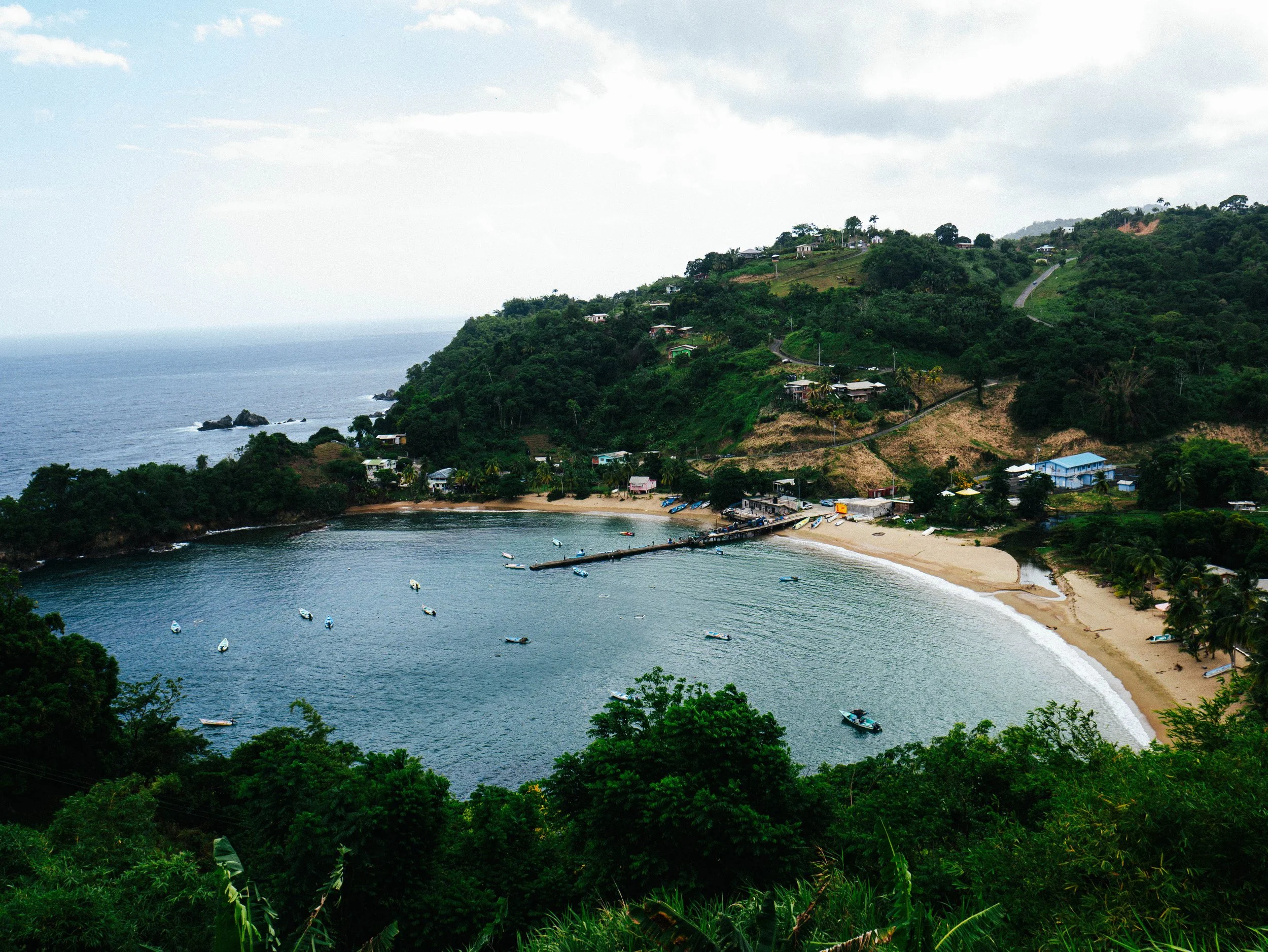 A coastal bay with several boats anchored in the water, a sandy beach, and a hillside with houses and lush greenery.