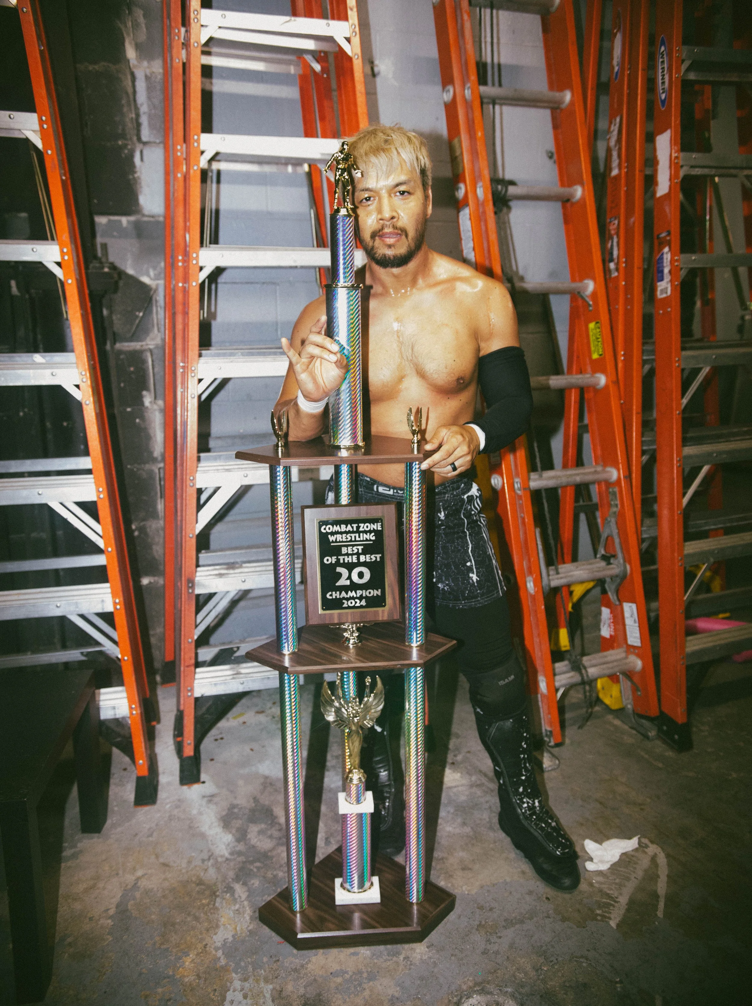 A shirtless wrestler with blonde hair and a black arm sleeve holding a large trophy in a backstage area with orange ladders in the background.