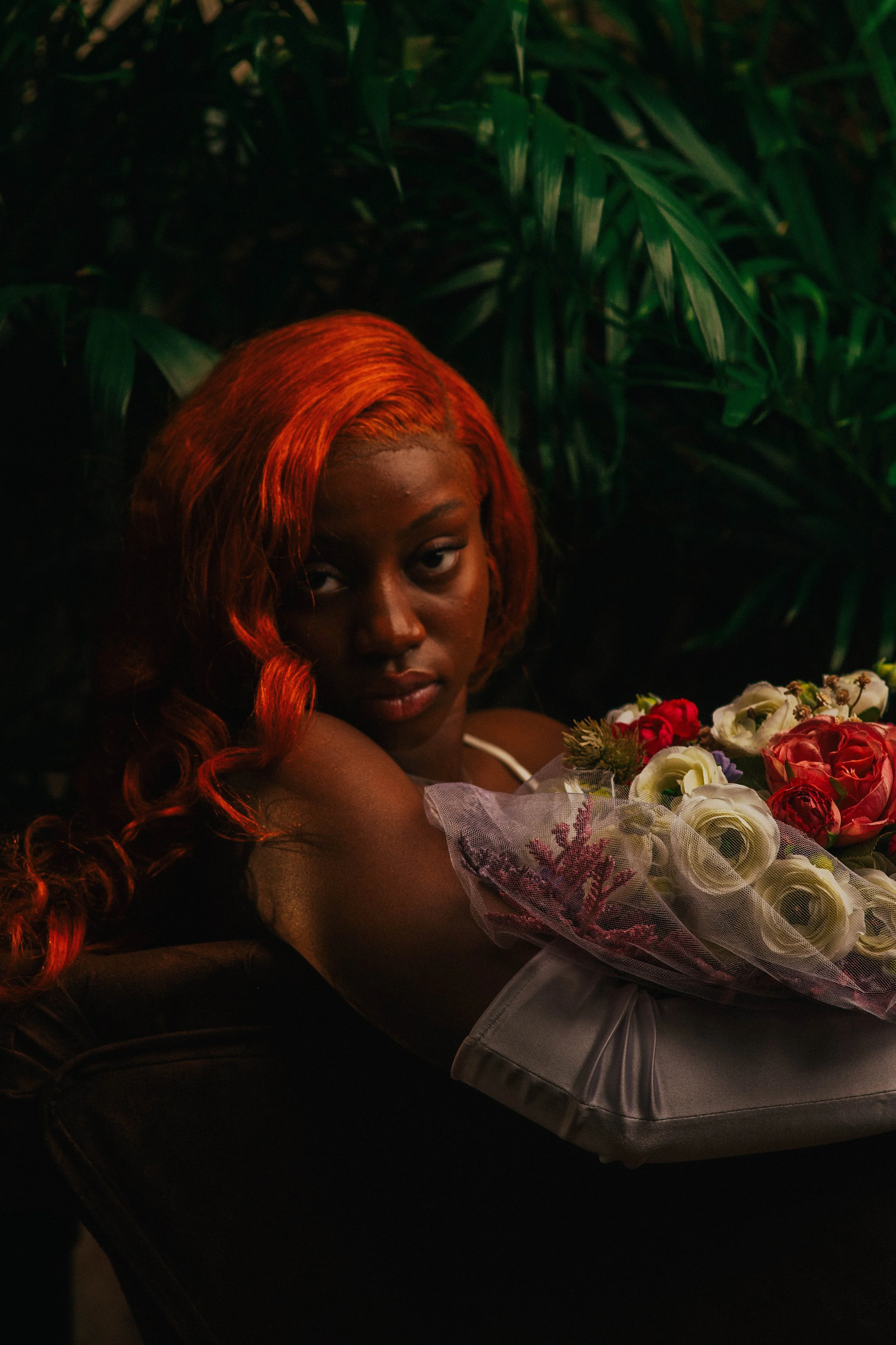 A woman with vibrant red hair resting her head on a chair, looking at the camera, holding a bouquet of white and red flowers, with lush green foliage in the background.