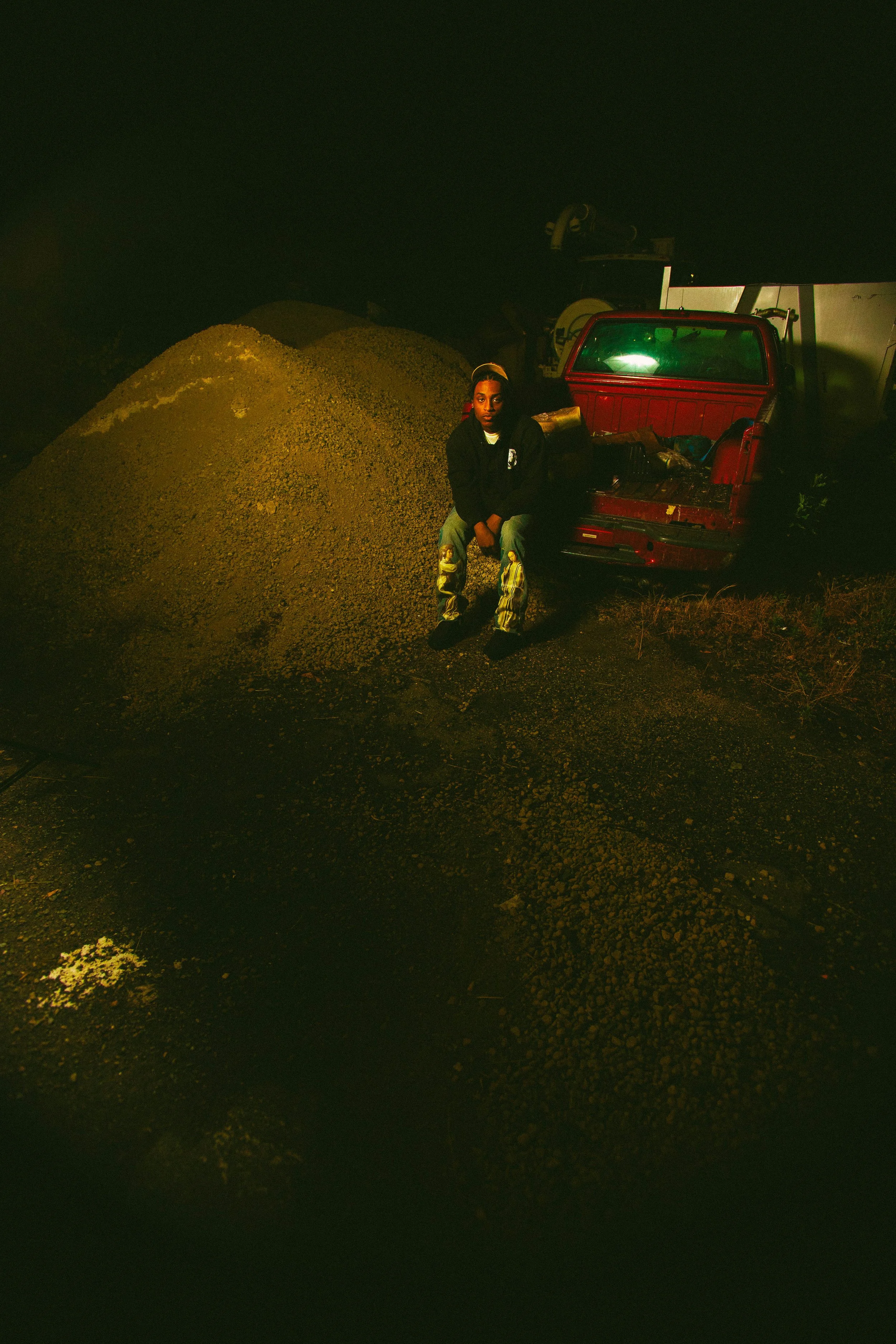 A person sitting on a small red truck bed next to a large pile of gravel or dirt at night with dark surroundings.