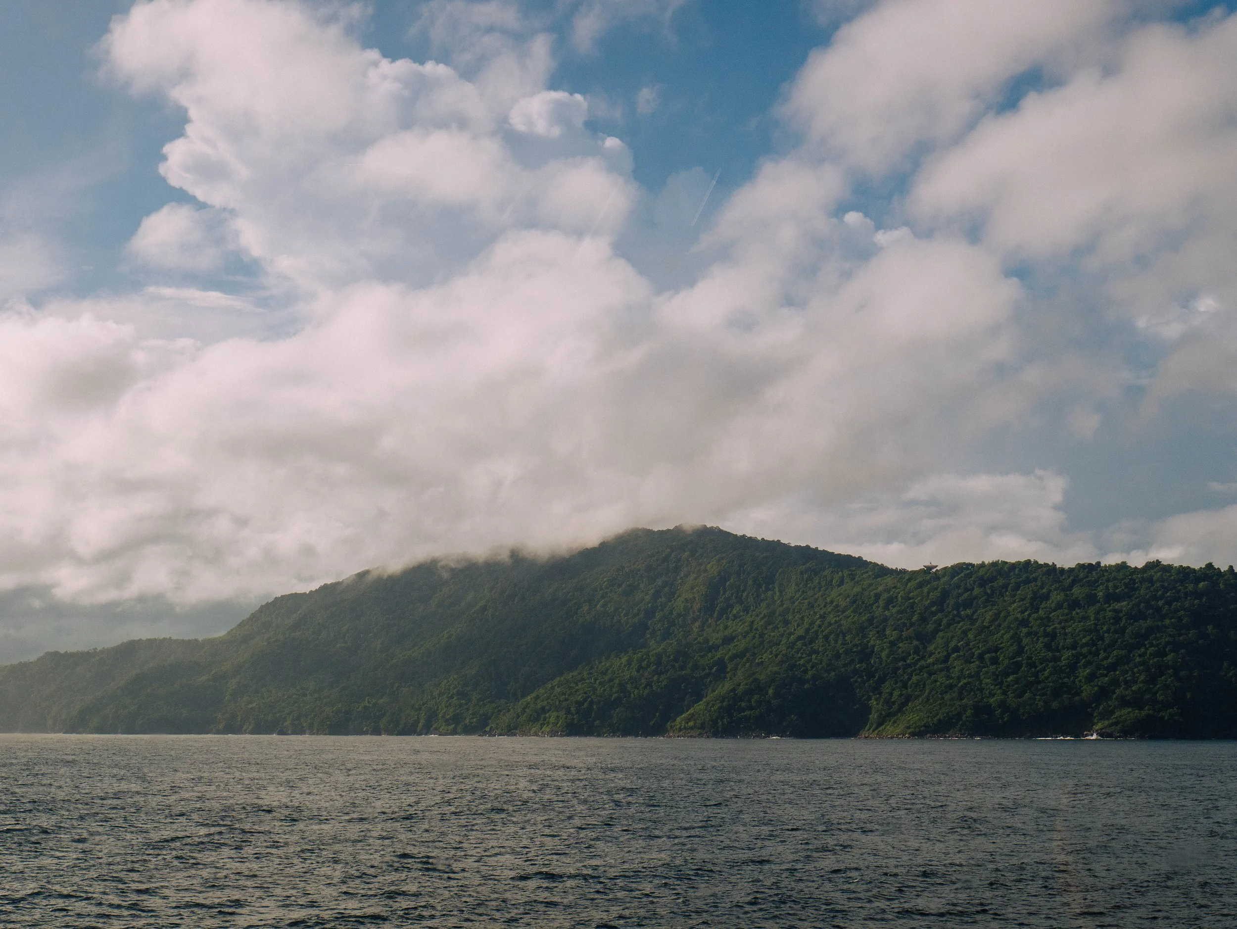 A large body of water in the foreground with green forested hills in the background under partly cloudy sky.