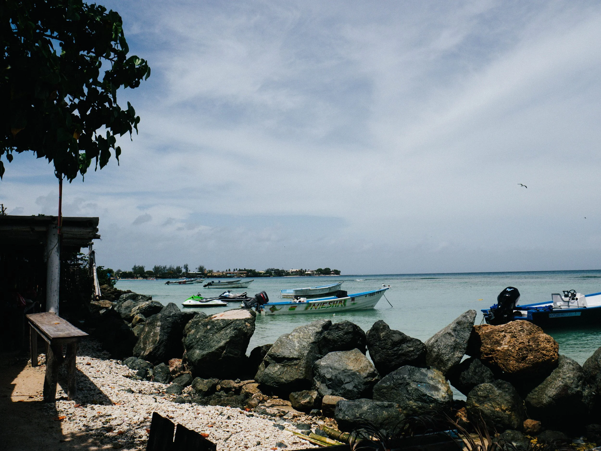 Boats anchored near a rocky shoreline on a cloudy day, with the ocean and land in the distance.