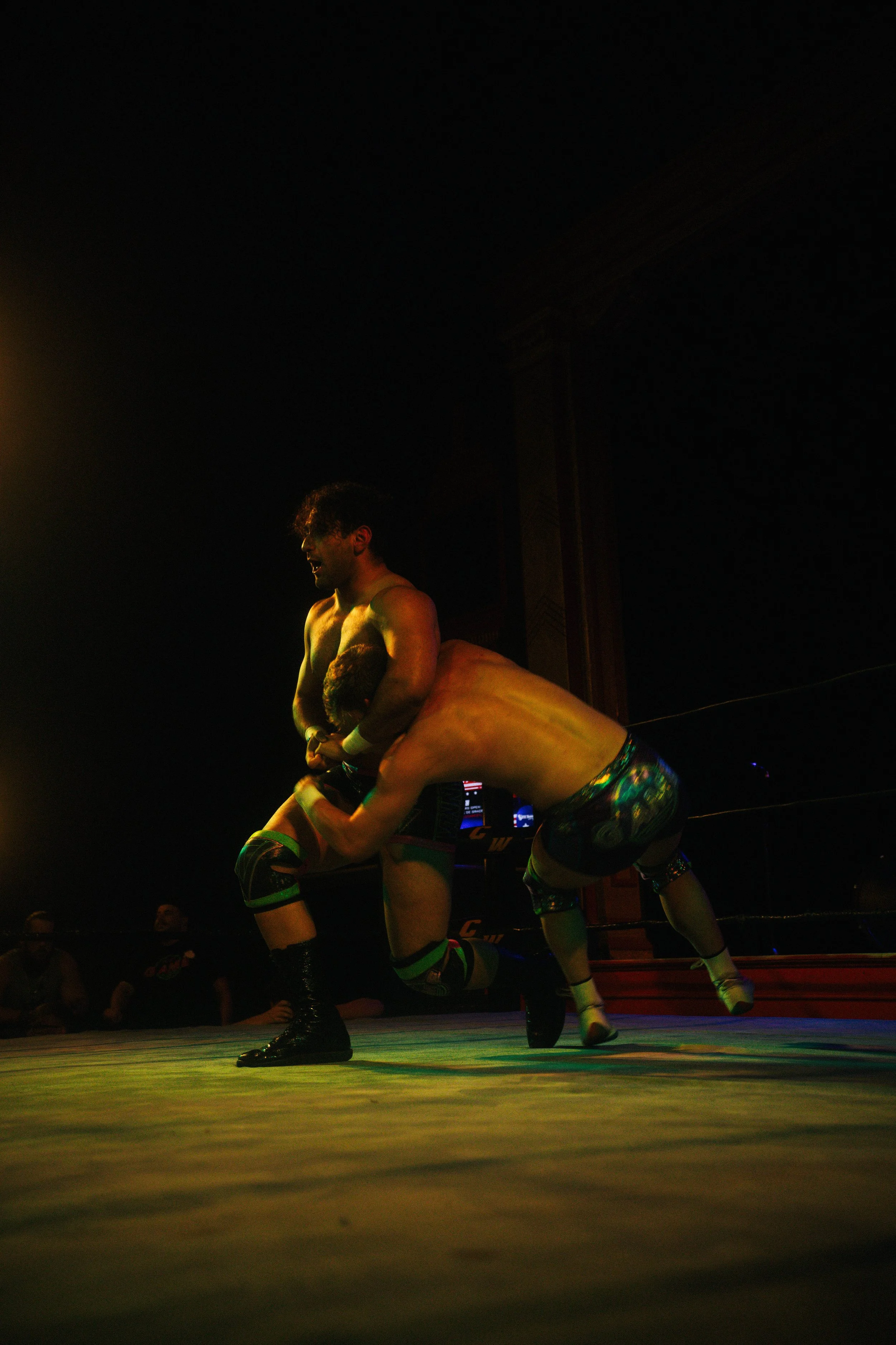 Two wrestlers in the midst of a wrestling match inside a ring, with one wrestler grasping the other's midsection as they perform a move, under dim lighting with a few audience members in the background.