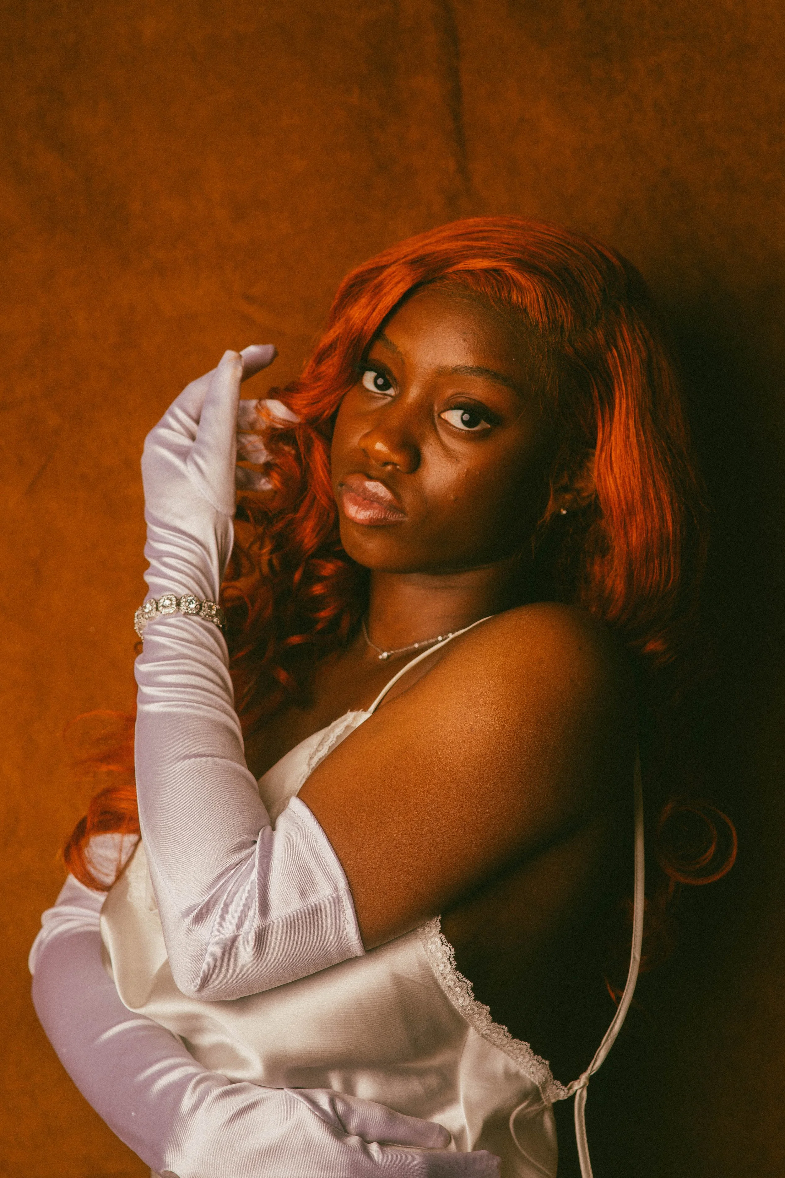 A woman with long, wavy red hair gazes at the camera while wearing white satin gloves, a delicate necklace, and lace-trimmed satin garment against a warm, textured background.