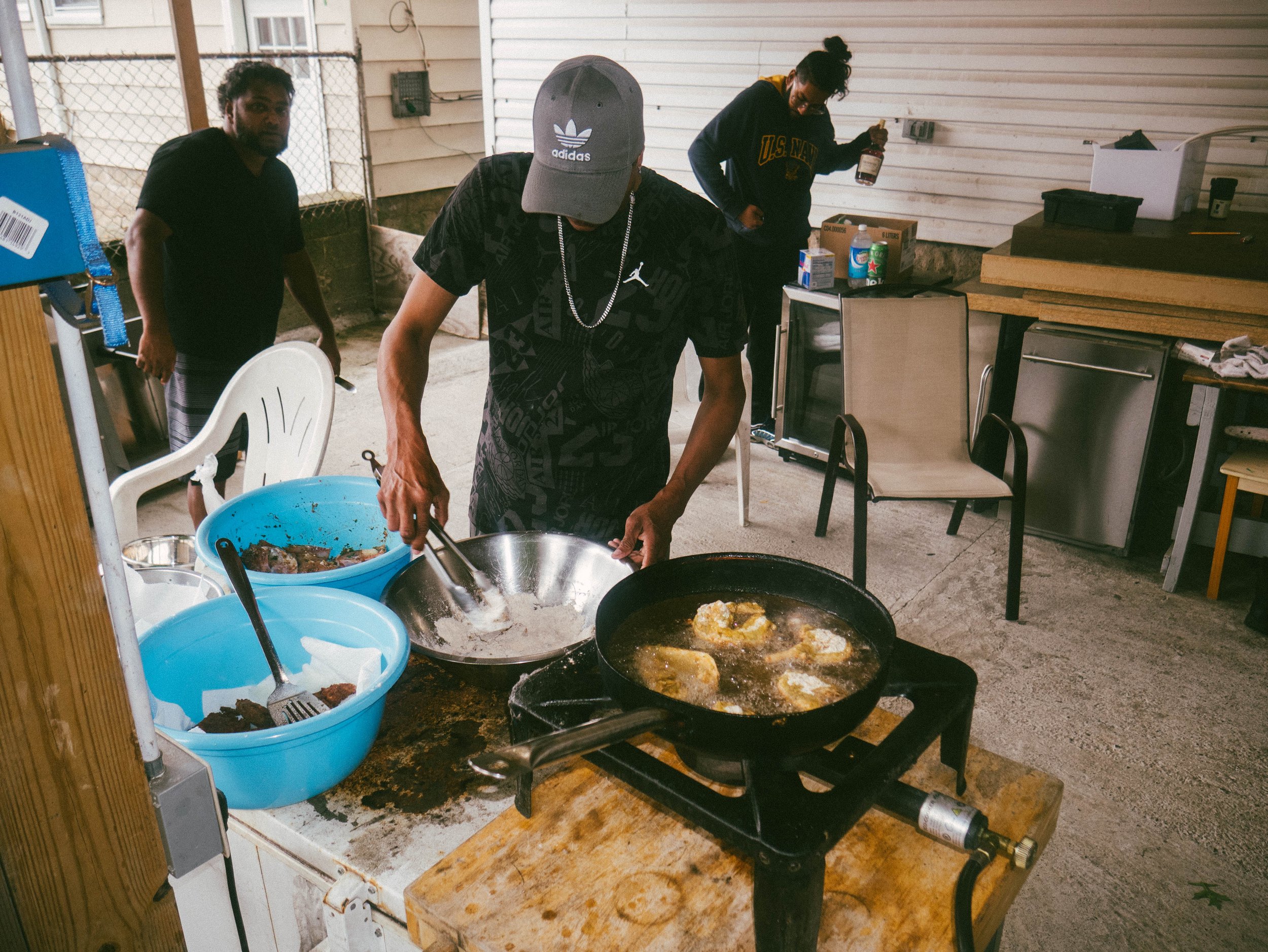Three men cooking dinner in a garage. One man is frying chicken in a skillet while another is seasoning food in a stainless steel mixing bowl. The third man is standing in the back near a table with drinks and food supplies.