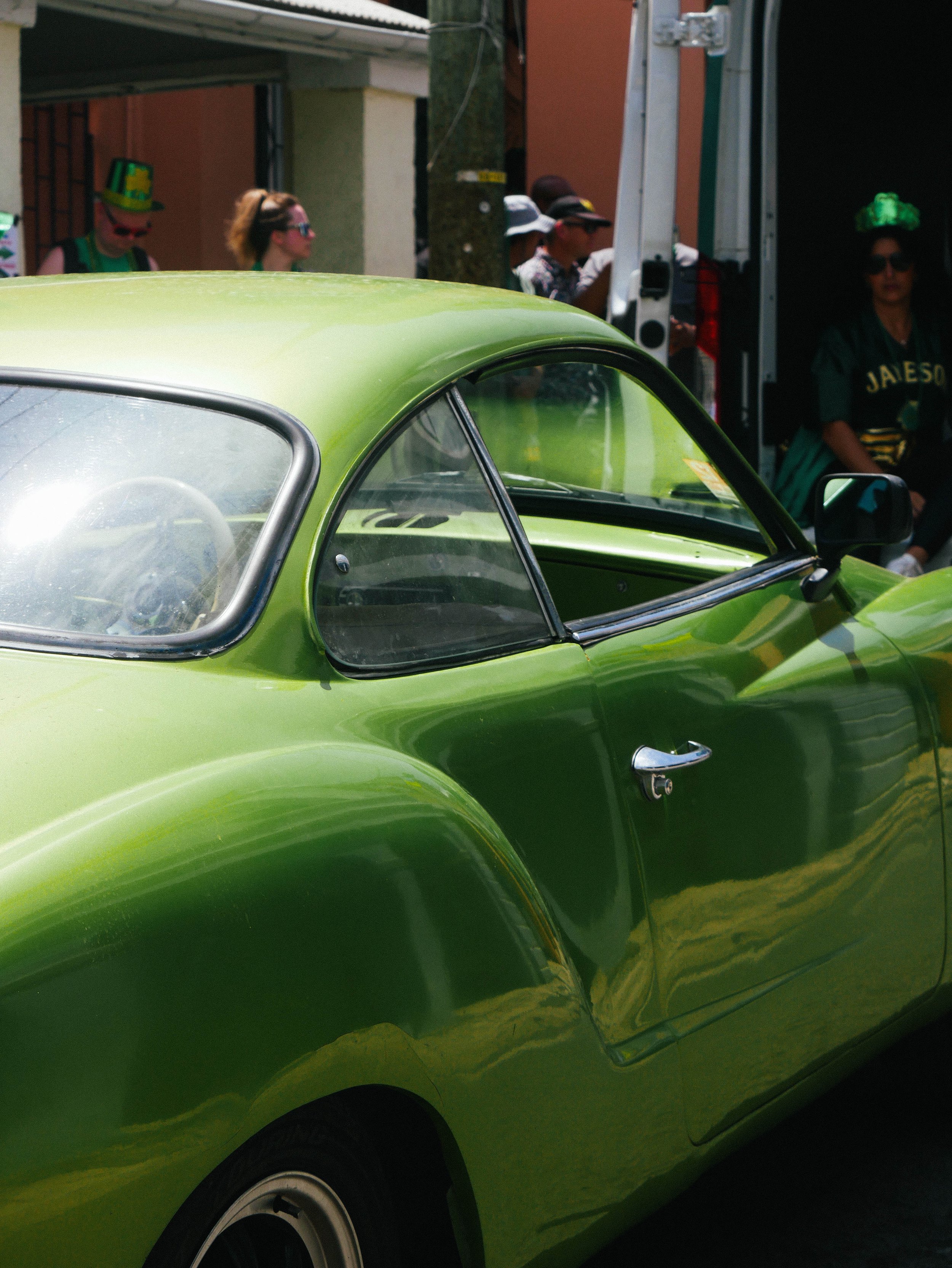 A bright green vintage car at an outdoor event, with people in the background wearing festive accessories and costumes.