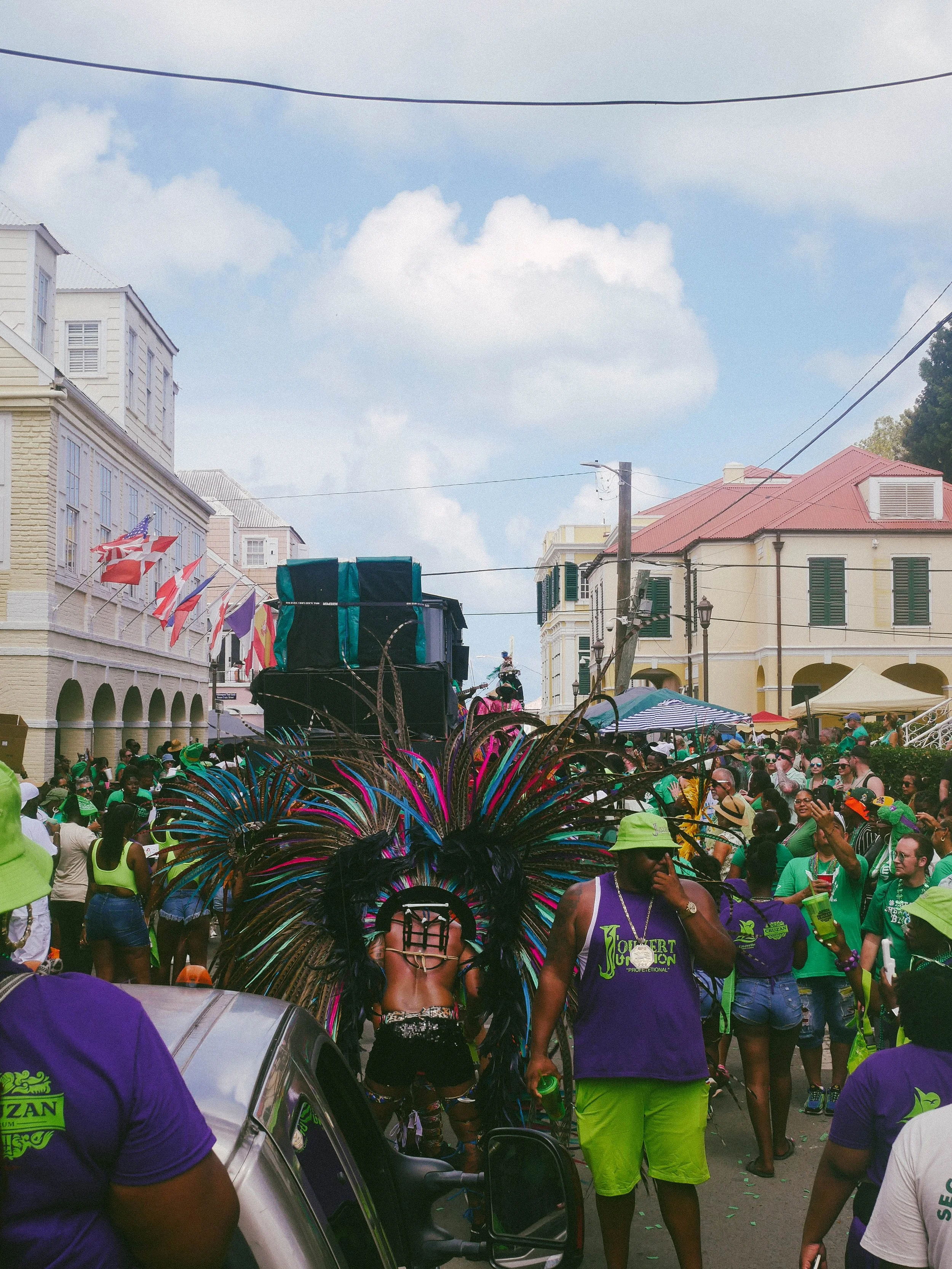 Crowd of people celebrating at a street parade with a performer in a vibrant feathered costume, surrounded by colorful tents and buildings.