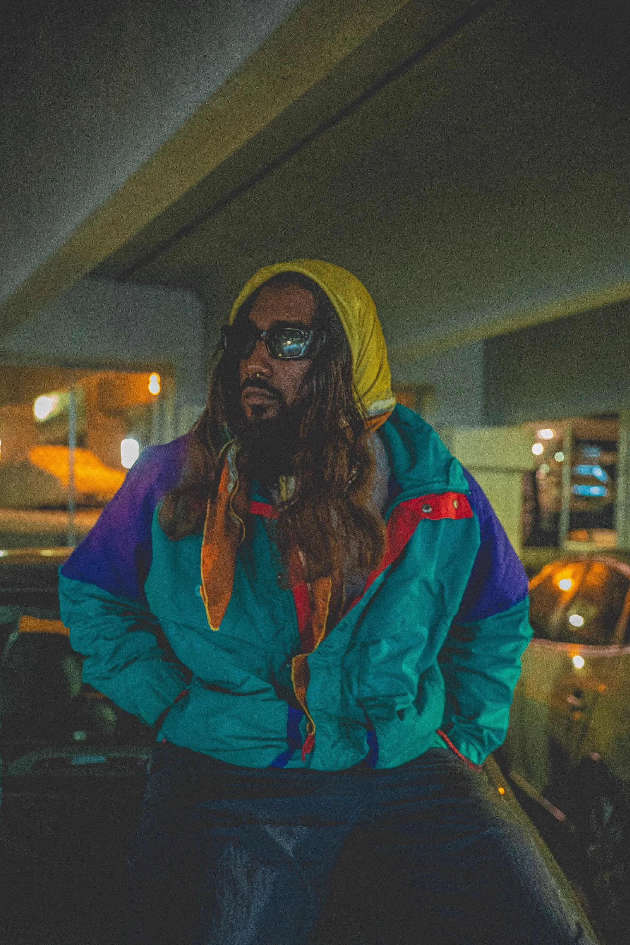 Person with long hair and sunglasses wearing a colorful windbreaker jacket sitting in a parking garage at night.
