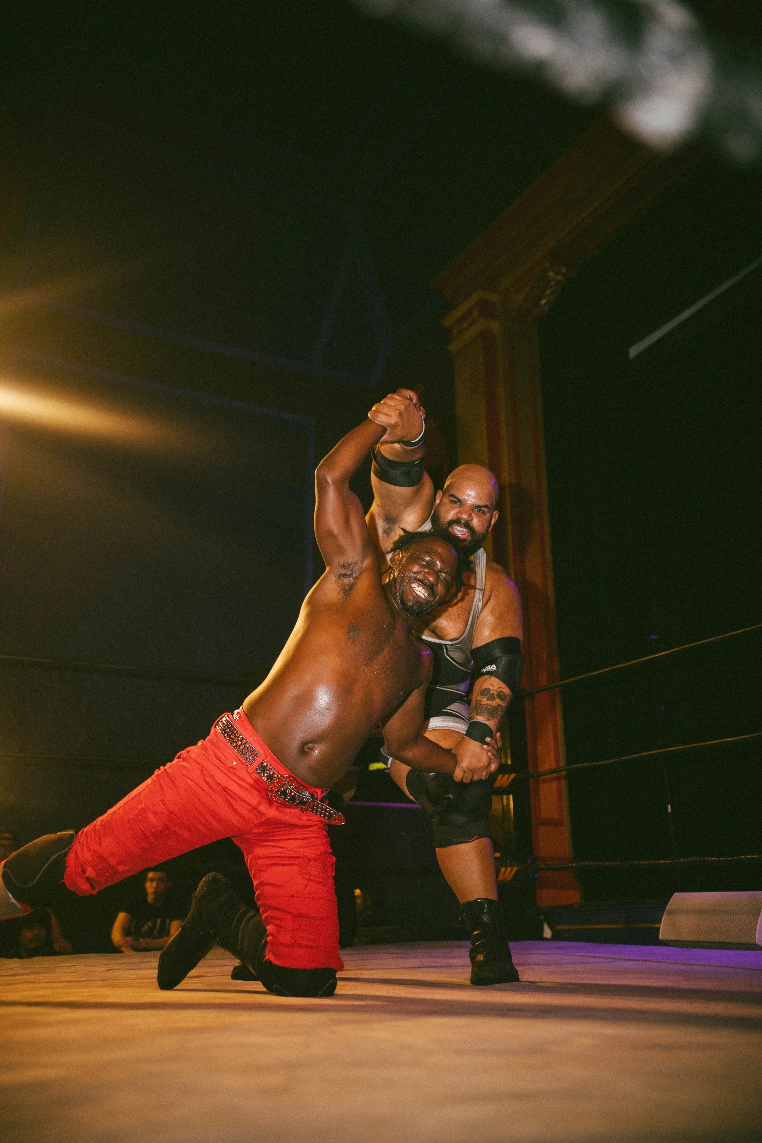 Two wrestlers celebrating victory inside a wrestling ring, with one on his knees and the other standing behind him, both smiling and showing strength.