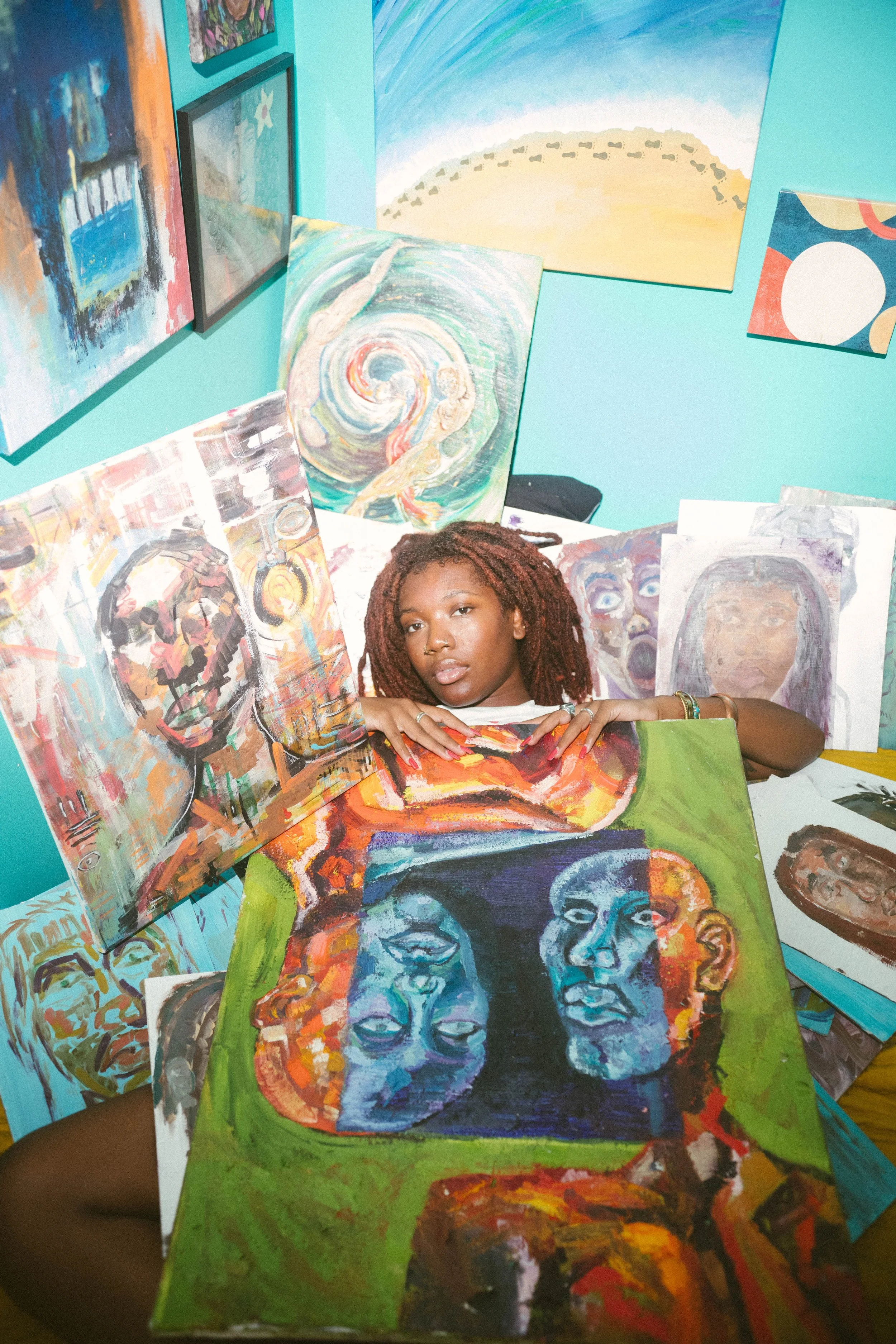 Young woman with red dreadlocks surrounded by colorful paintings, holding a painting of multiple faces with various expressions at an art studio with blue walls and additional artwork on the walls.