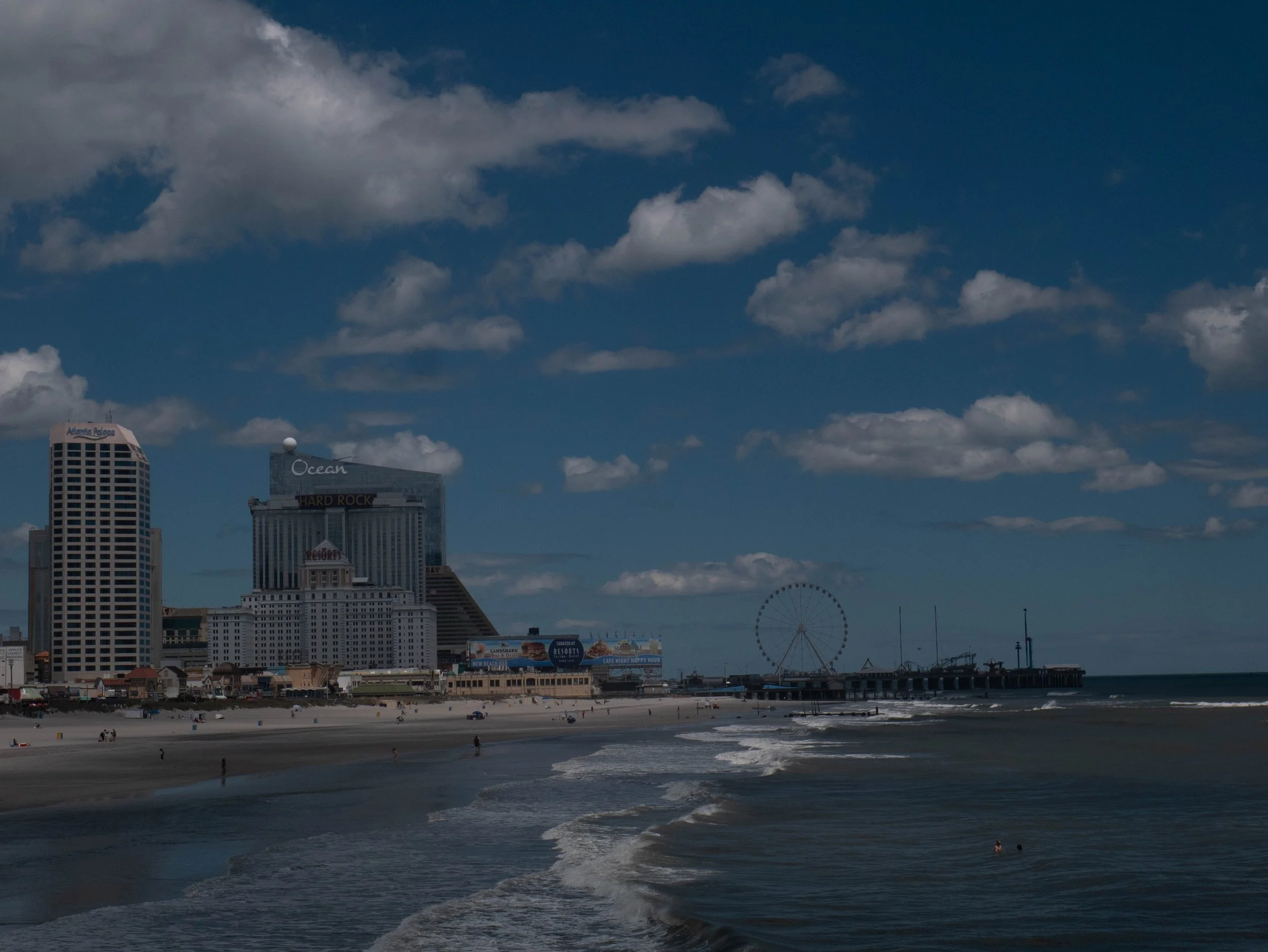 Bright city skyline with tall buildings, a beach with people, and a Ferris wheel in the distance under a partly cloudy sky.