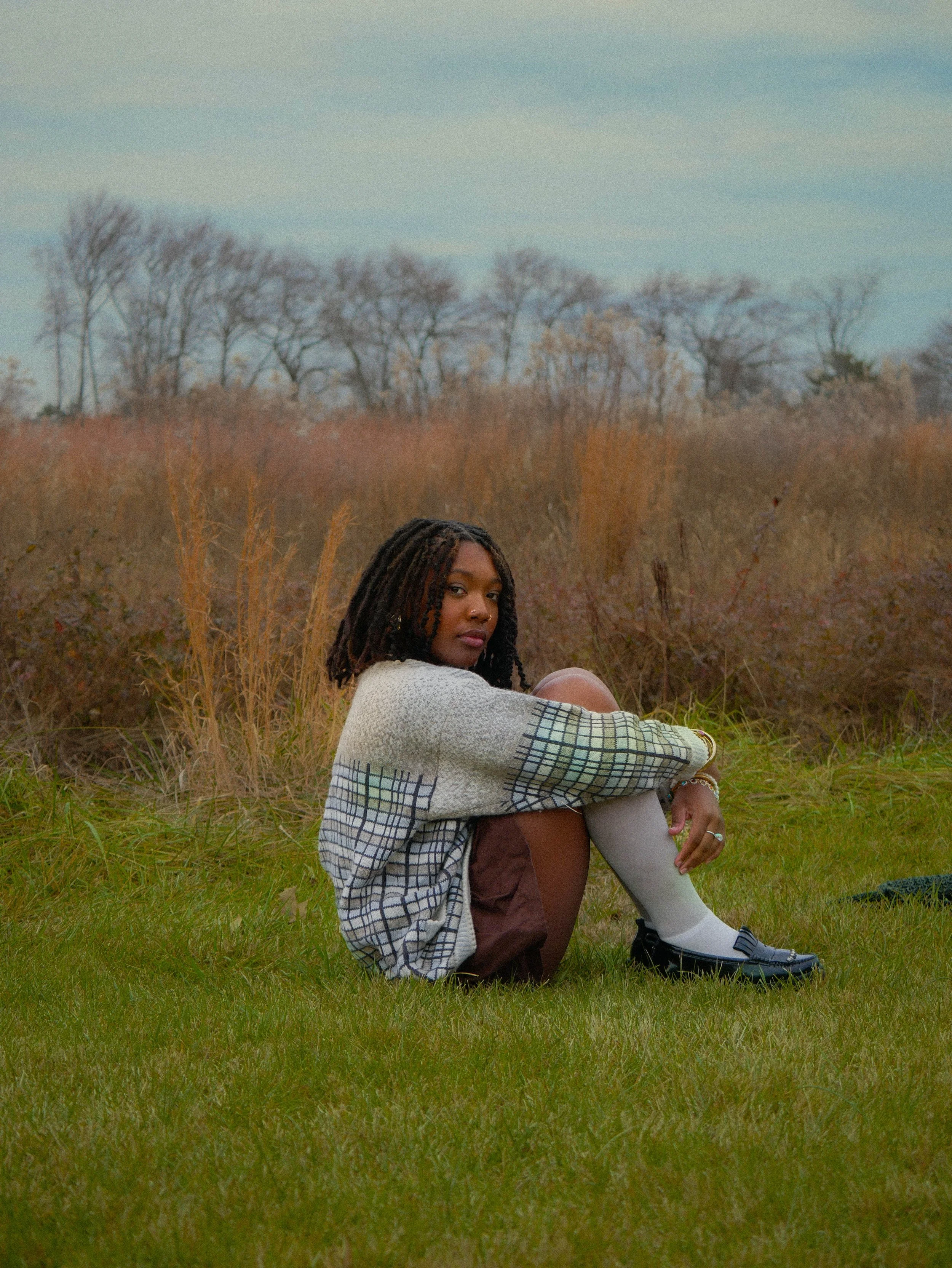 Young woman with dreadlocks sitting on grass in a field during fall, wearing a plaid sweater, brown shorts, and white knee-high socks with black loafers, with trees in the background under a partly cloudy sky.