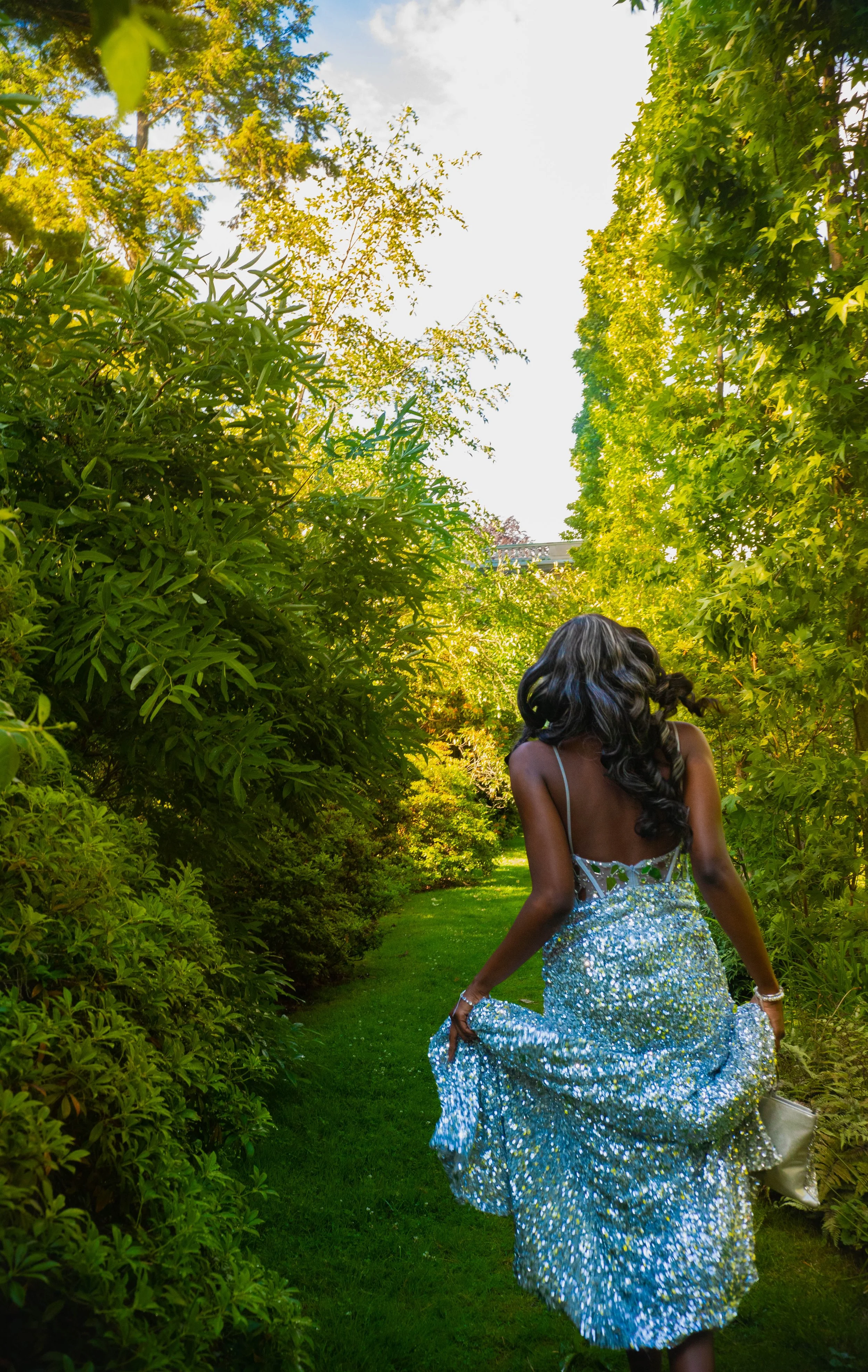 A woman walking through a lush green garden at sunset, wearing a sparkly silver dress and holding it up slightly to show her shoes and accessories.