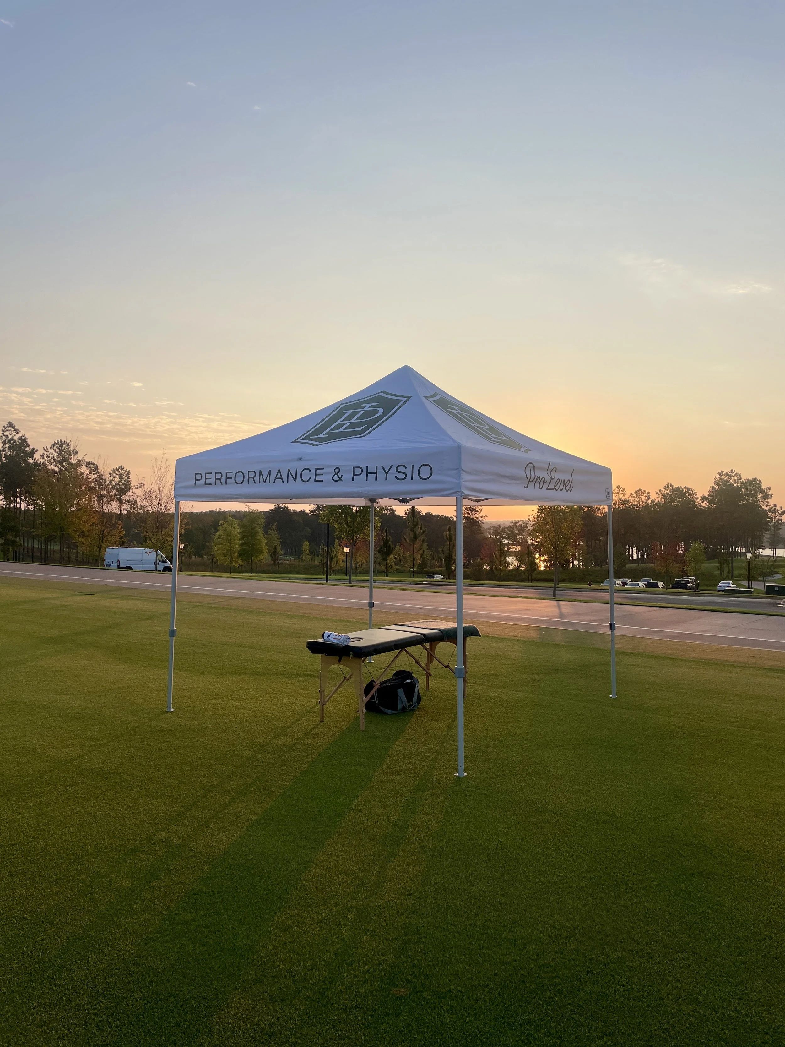 A white canopy tent with the words 'Performance & Physio' and 'Pro Level' set up on a grassy area during sunset, with a park and parking lot in the background.