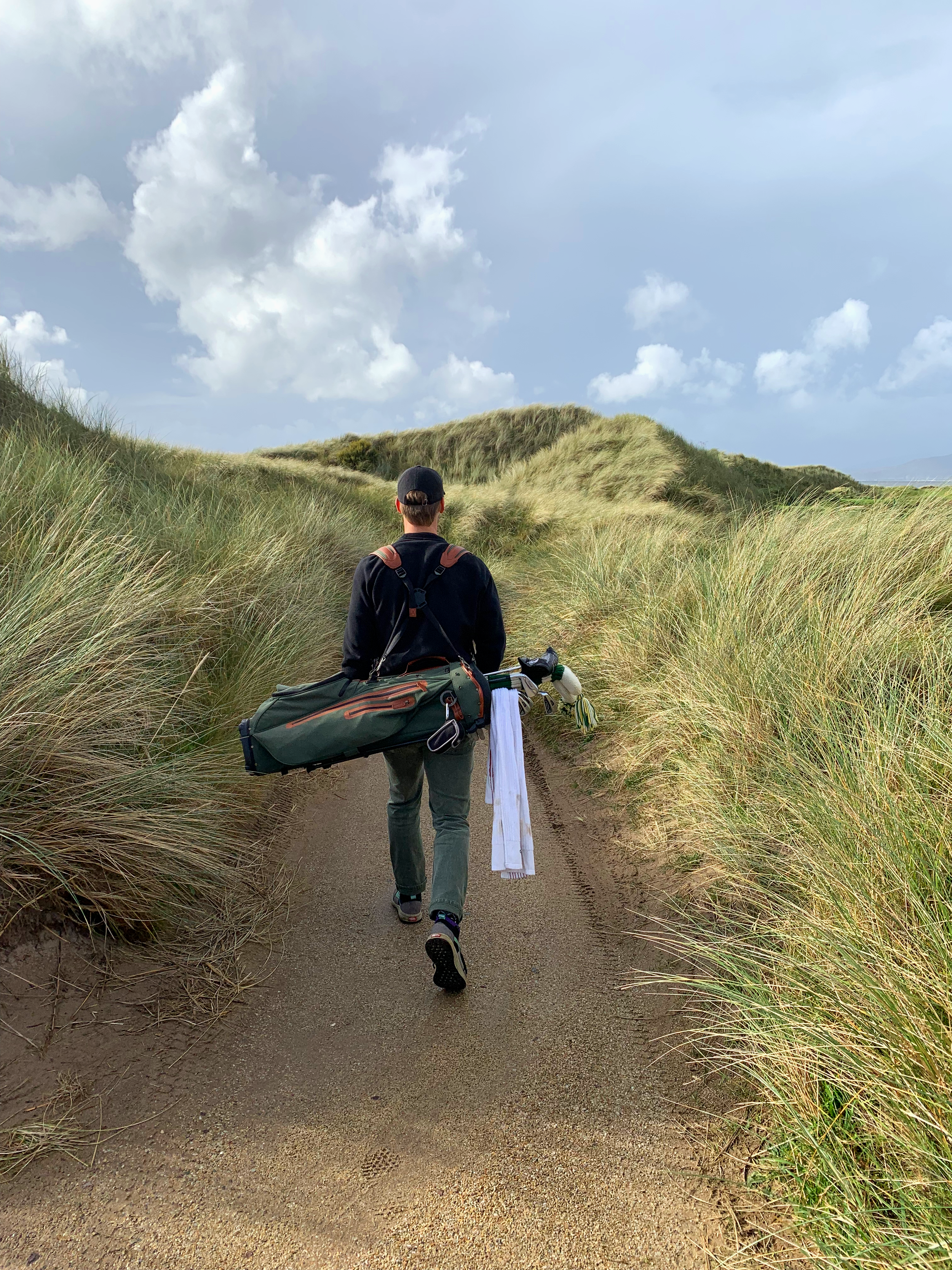 A person wearing a black hoodie, gray pants, and a black cap walking on a sandy trail through tall green grass with a backpack and a towel hanging from it. The sky is partly cloudy.