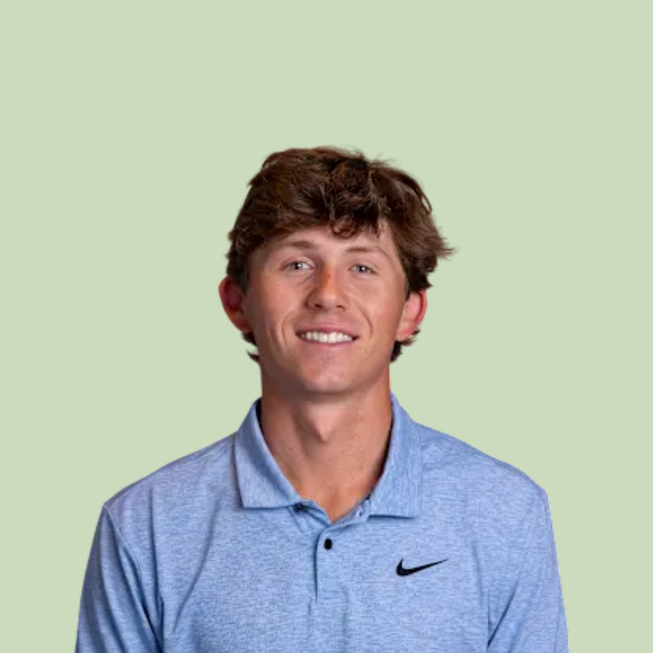 Close-up of a young man with curly brown hair, blue eyes, and light skin, wearing a light gray Nike polo shirt, smiling against a pale green background.