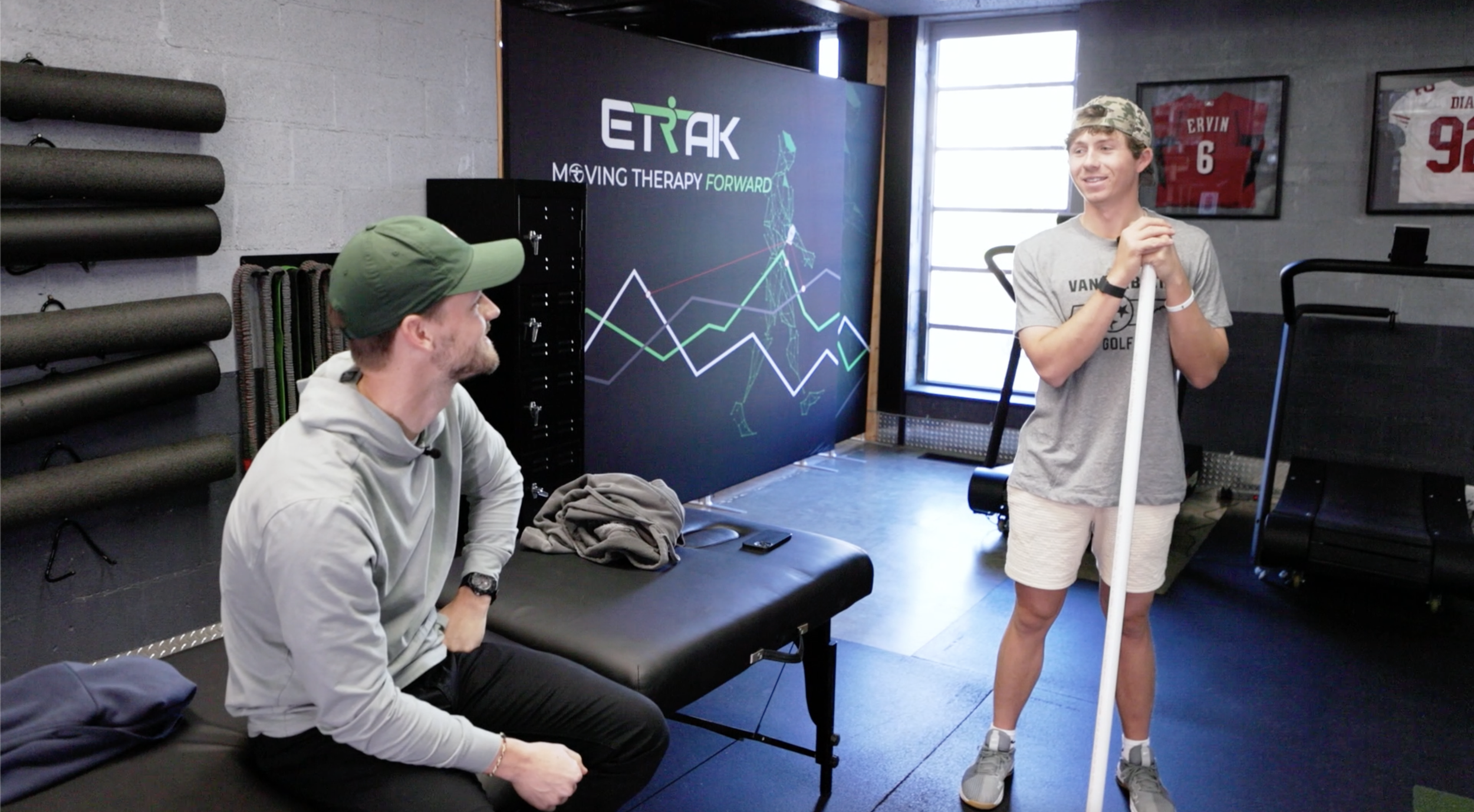 Two men are having a conversation in a fitness or therapy center. One man is seated on a bench wearing a gray shirt and green cap, while the other man stands holding a white pole, wearing a gray T-shirt, shorts, and a camouflage cap. The room has black foam rollers on the wall, framed sports jerseys, and a digital display with a running figure and graph lines, indicating a focus on movement therapy or sports training.