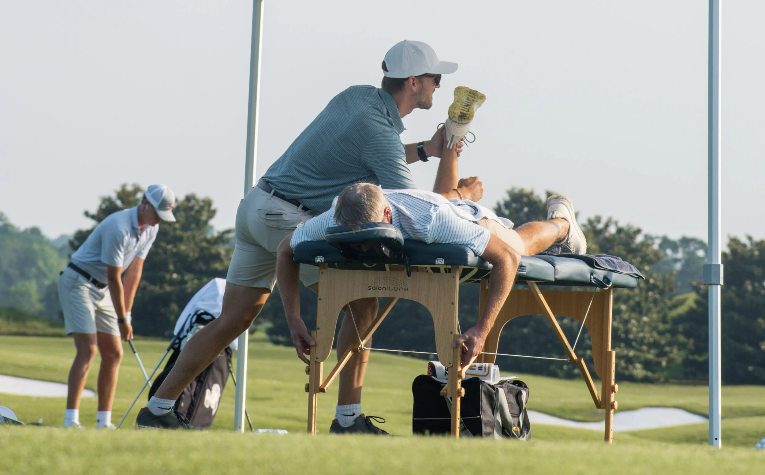 A man receiving a medical or sports massage on his back on a massage table outdoors at a golf course, with golfers practicing in the background.