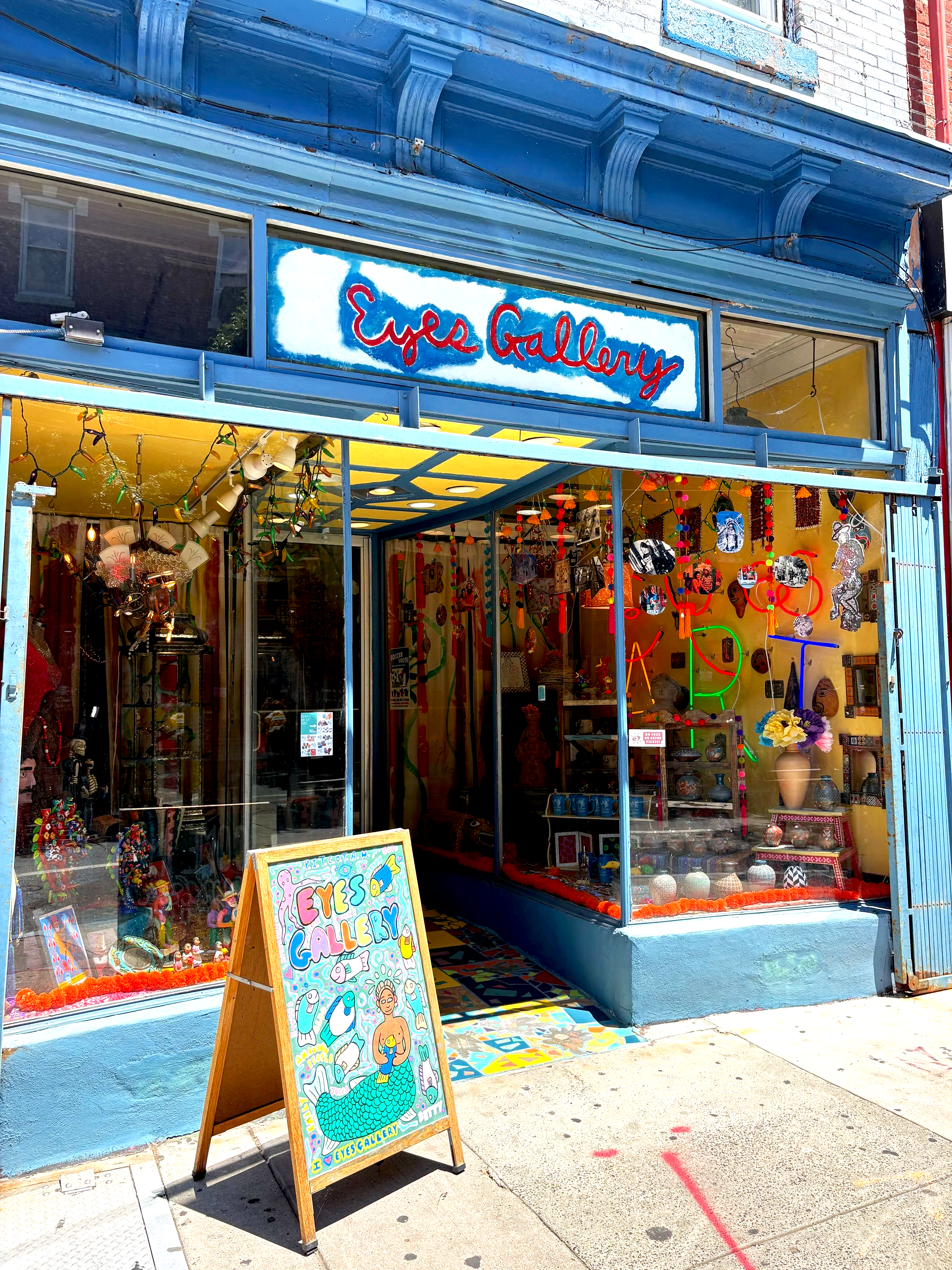 The storefront of Eye's Gallery on South Street in Philadelphia, PA. The display window shows various colorful art and decorative items, including necklaces and small sculptures. A colorful chalkboard sign outside promotes the shop's name and art.