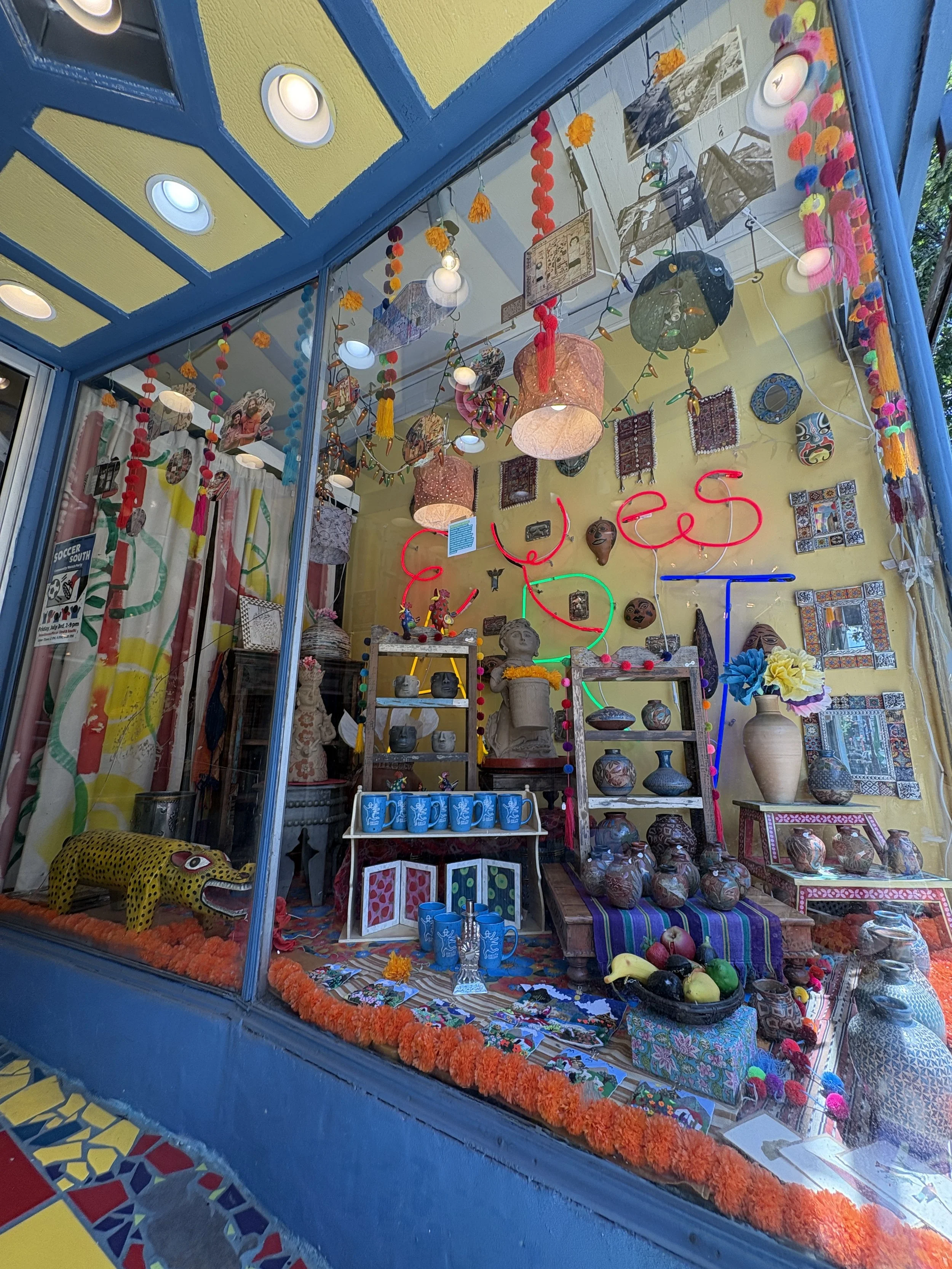Colorful display of Mexican crafts and decor in the shop window of Eye's Gallery. Featuring pottery, masks, textiles, and papel picado, with vibrant hanging decorations and framed pictures.