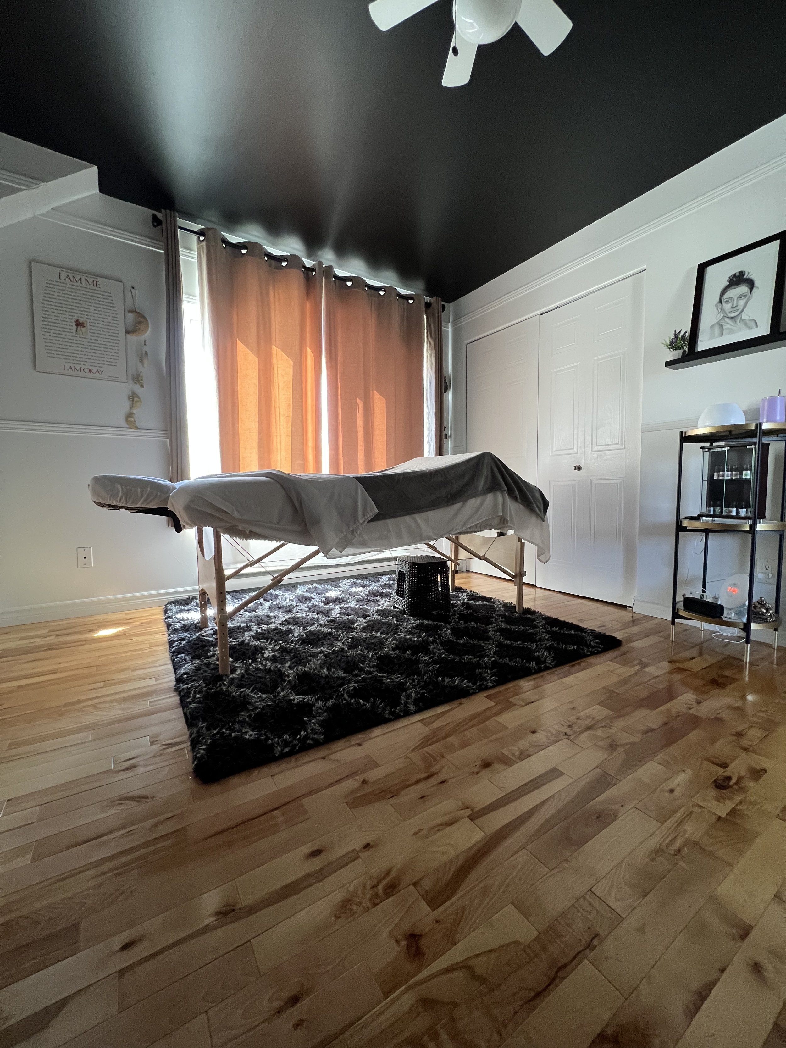 Massage room with a massage table, hardwood floor, black rug, curtains, and a ceiling fan.