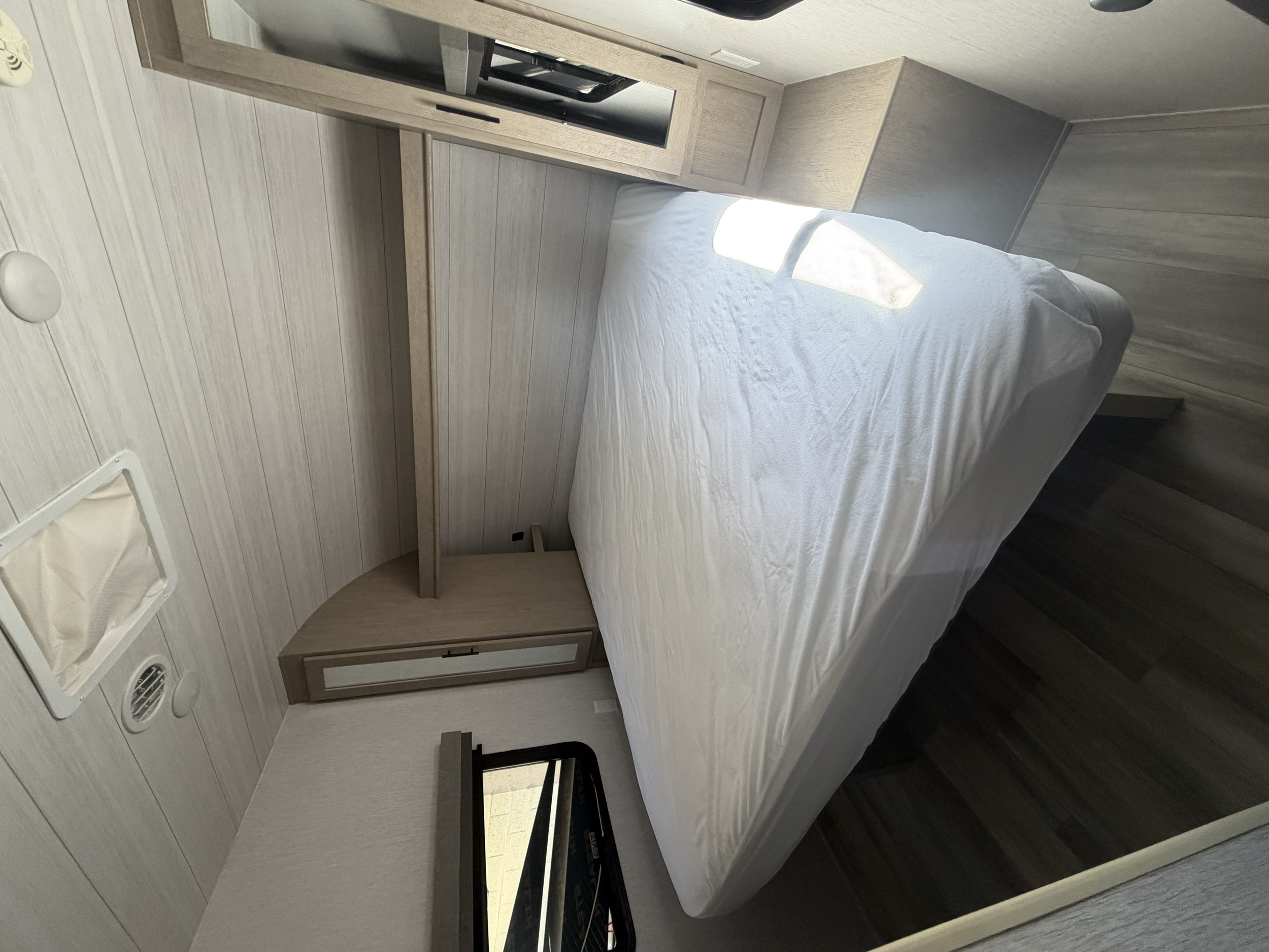 Interior of a small camper or RV bedroom with a bed, wooden cabinetry, a drain vent, and a TV mounted above the headboard.