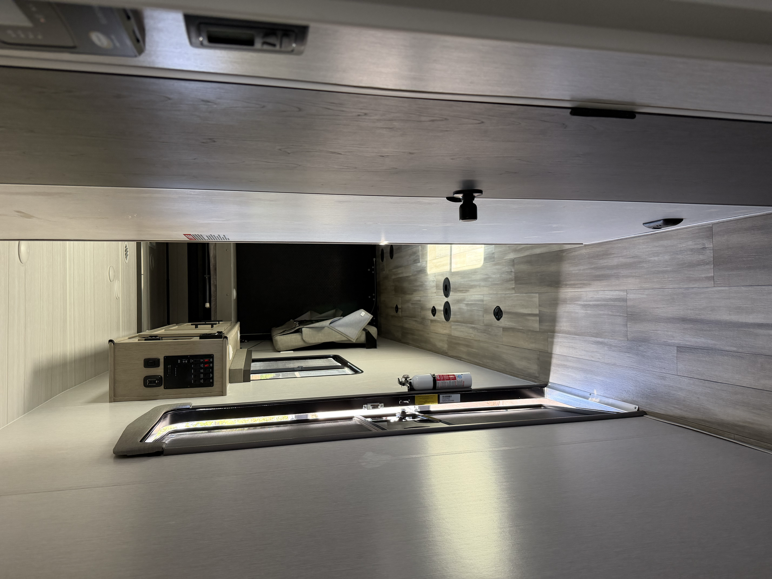 Interior of a modern RV or camper with wood paneling, a small kitchen area, and a seat with a blanket. The flooring is wood-look, and there are various fixtures including a fire extinguisher on the wall and circular vents on the floor.